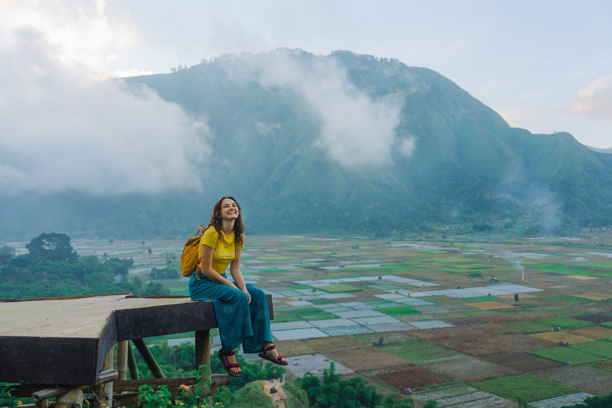 A smiling person in yellow sits on a ledge, surrounded by lush fields and mountains shrouded in clouds.
