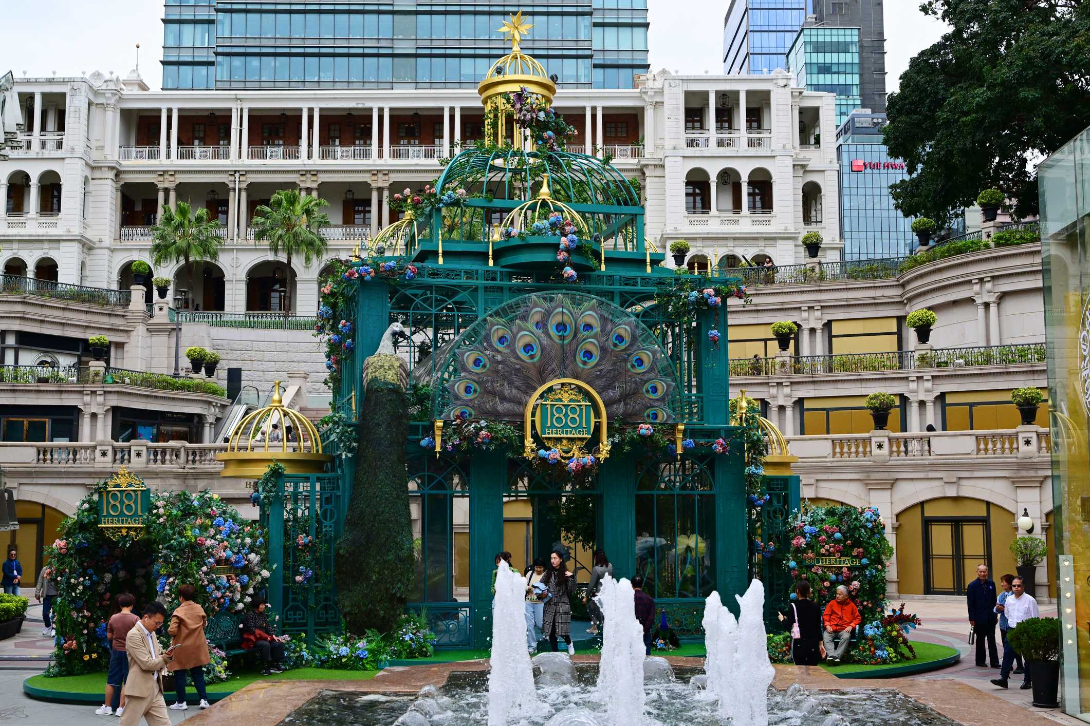 A colorful structure with peacock design and fountains, surrounded by greenery and buildings in a bustling plaza.