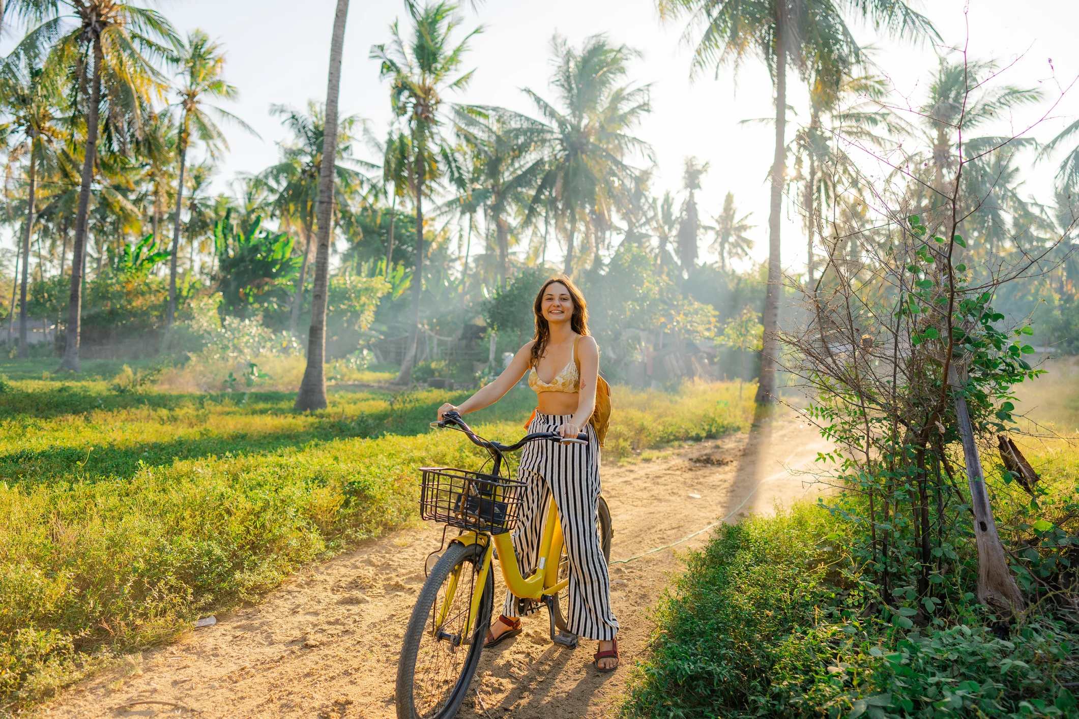 A woman rides a yellow bicycle on a dirt path surrounded by greenery and palm trees, with sunlight filtering through.