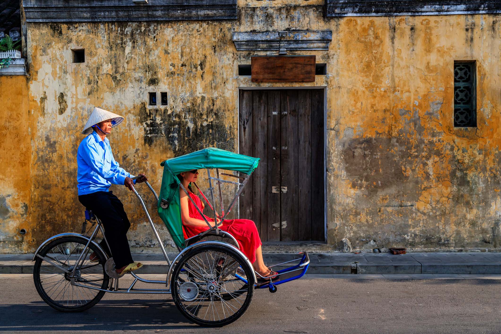 A rickshaw driver in a conical hat transports a woman in a red dress against a weathered yellow wall.