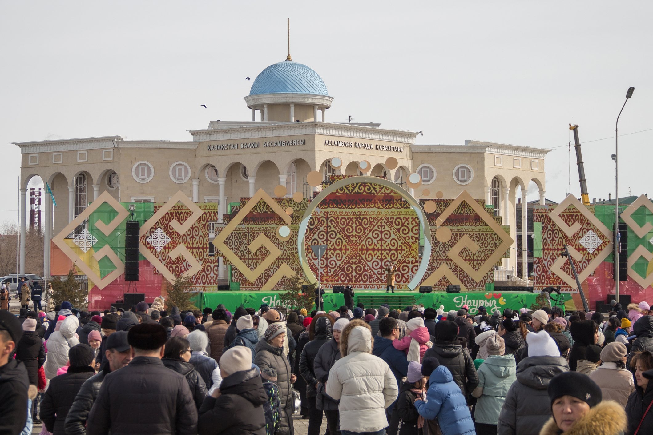 Crowd gathered in front of a large building with decorative patterns for a public event outdoors on a clear day.