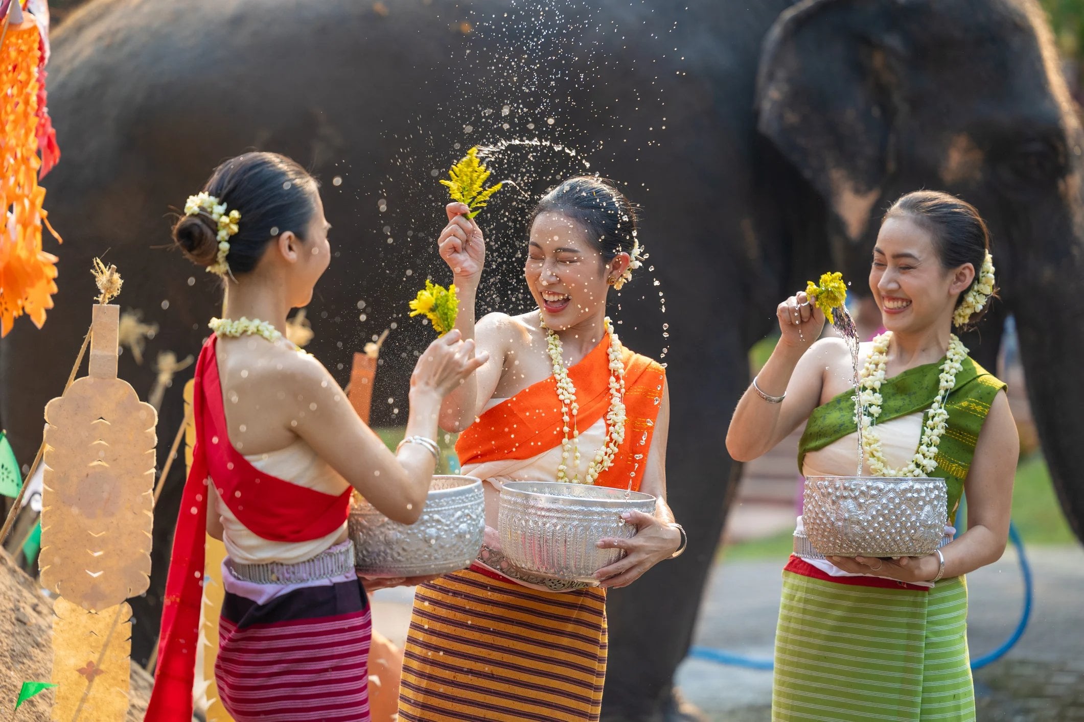 Three women in colorful traditional outfits joyfully splash water while celebrating with an elephant in the background.