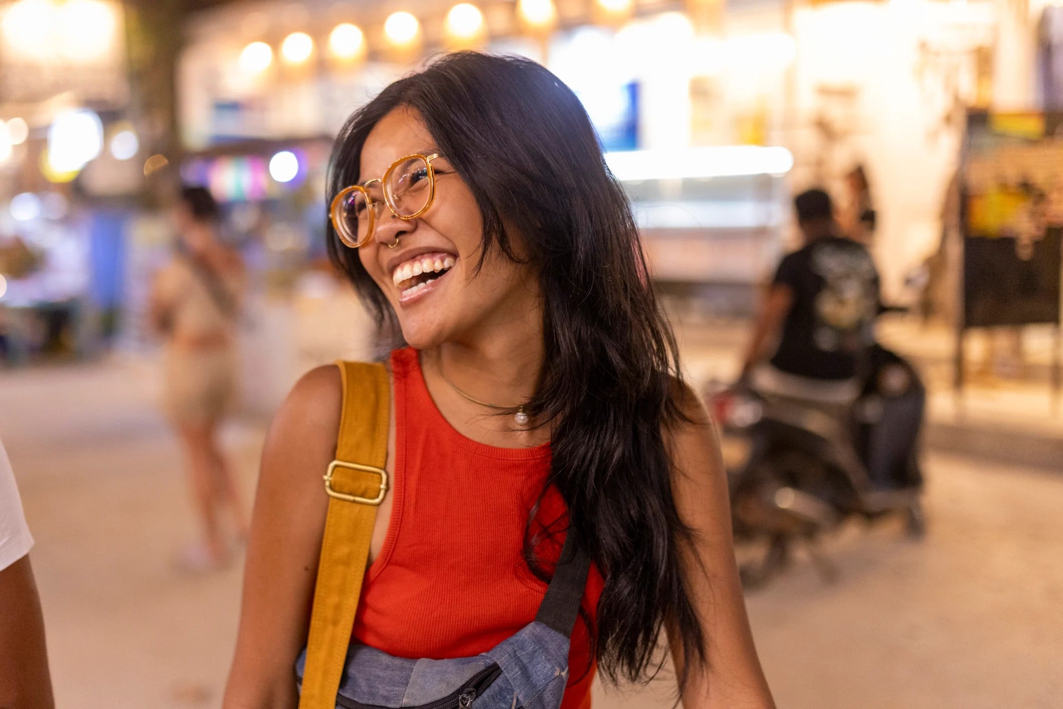 Smiling woman with long black hair, wearing glasses and a red shirt, standing in a bustling outdoor setting.