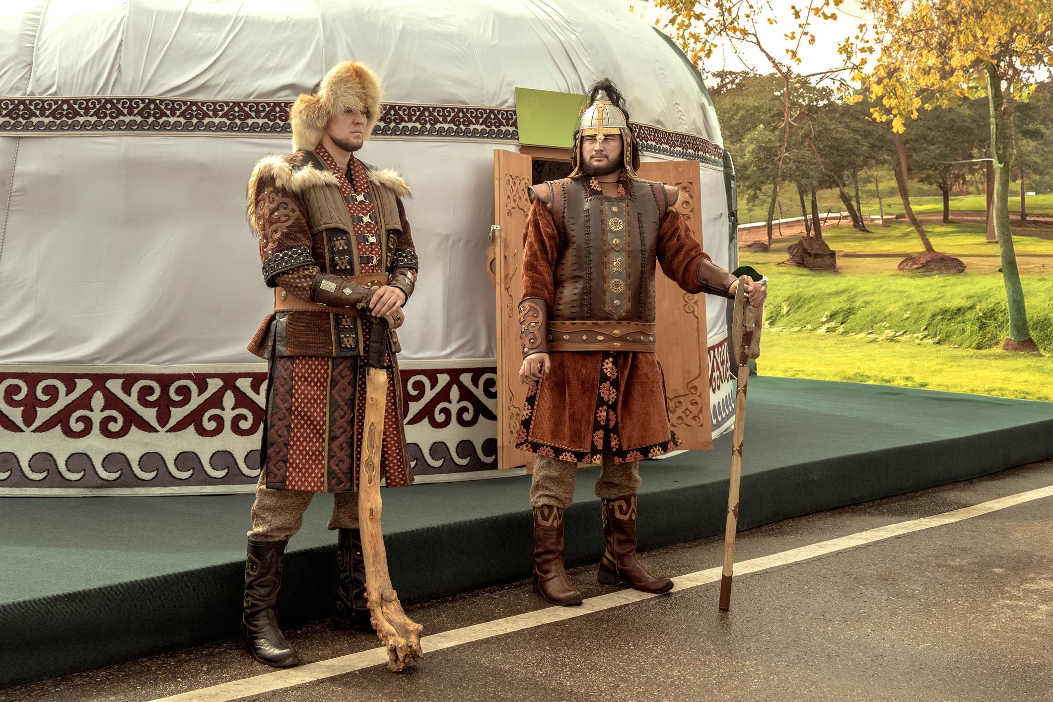 Two men in traditional clothing stand outside a yurt, holding staffs, with a green landscape in the background.