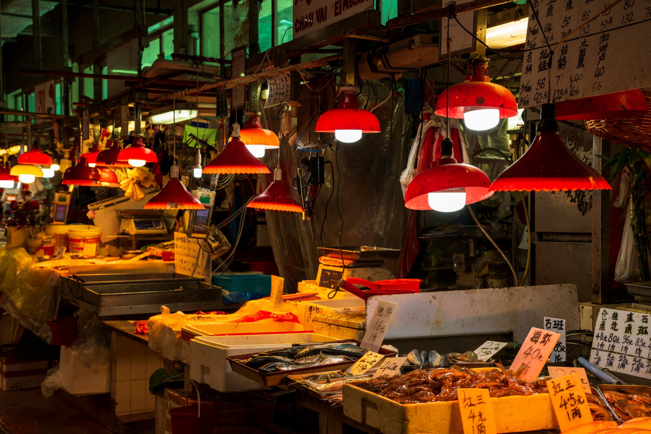 A bustling market stall with red lamps illuminating fresh seafood and produce, surrounded by vibrant signboards.