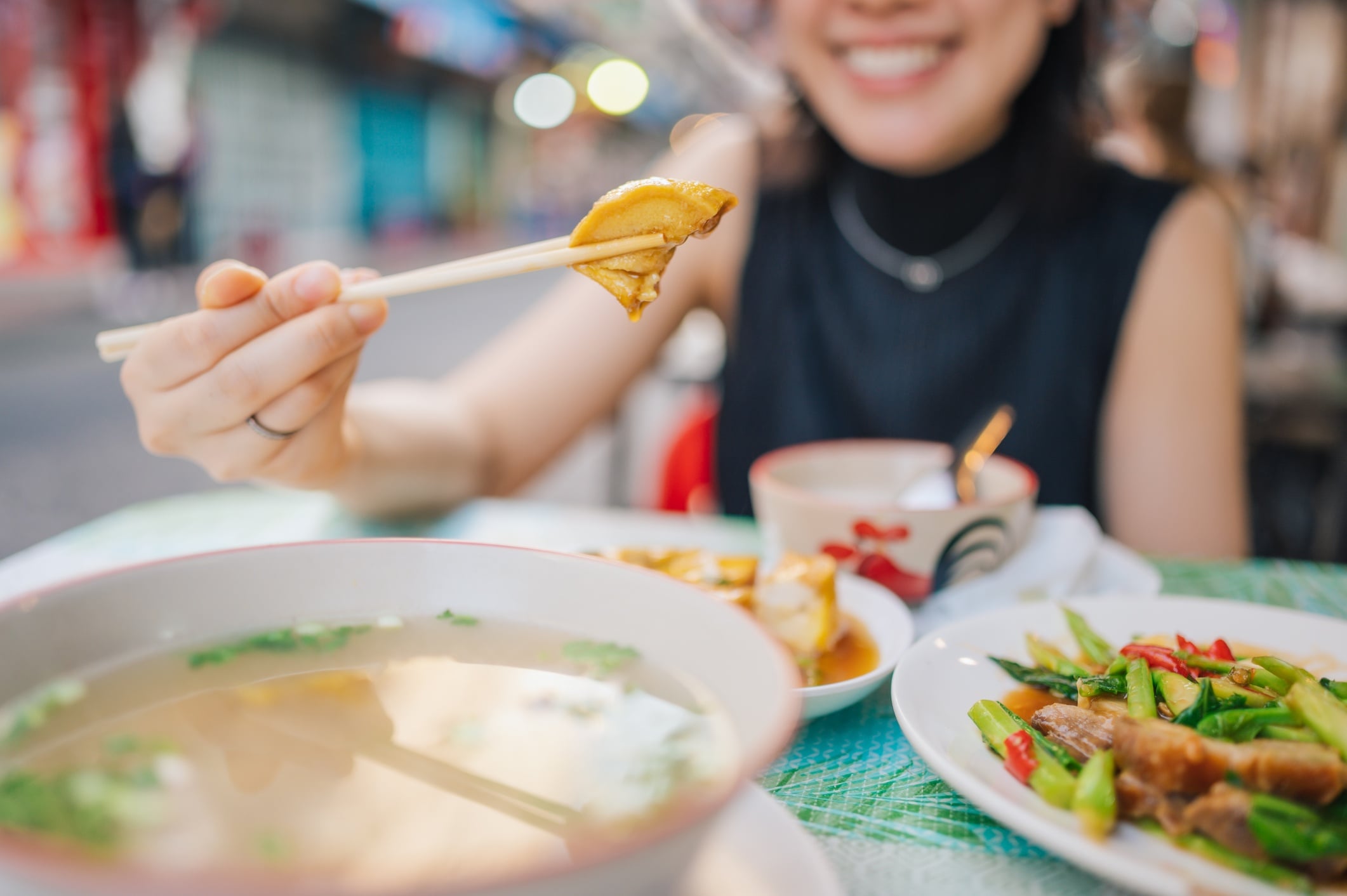 A person holds a piece of food with chopsticks, smiling at a table with a bowl of soup and colorful dishes.