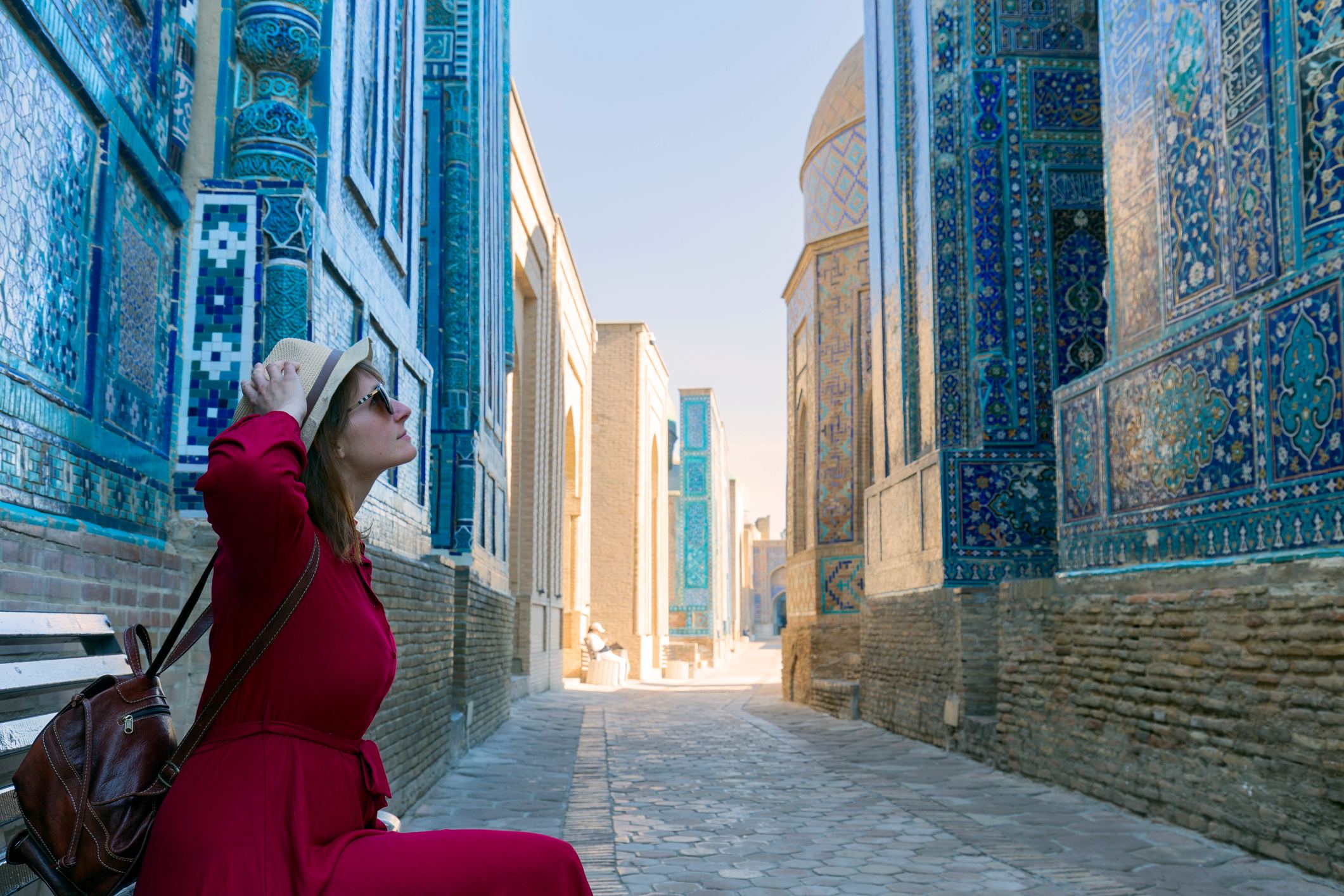 A woman in a red dress and hat sits on a bench in a narrow, blue-tiled alleyway, looking up at the architecture.