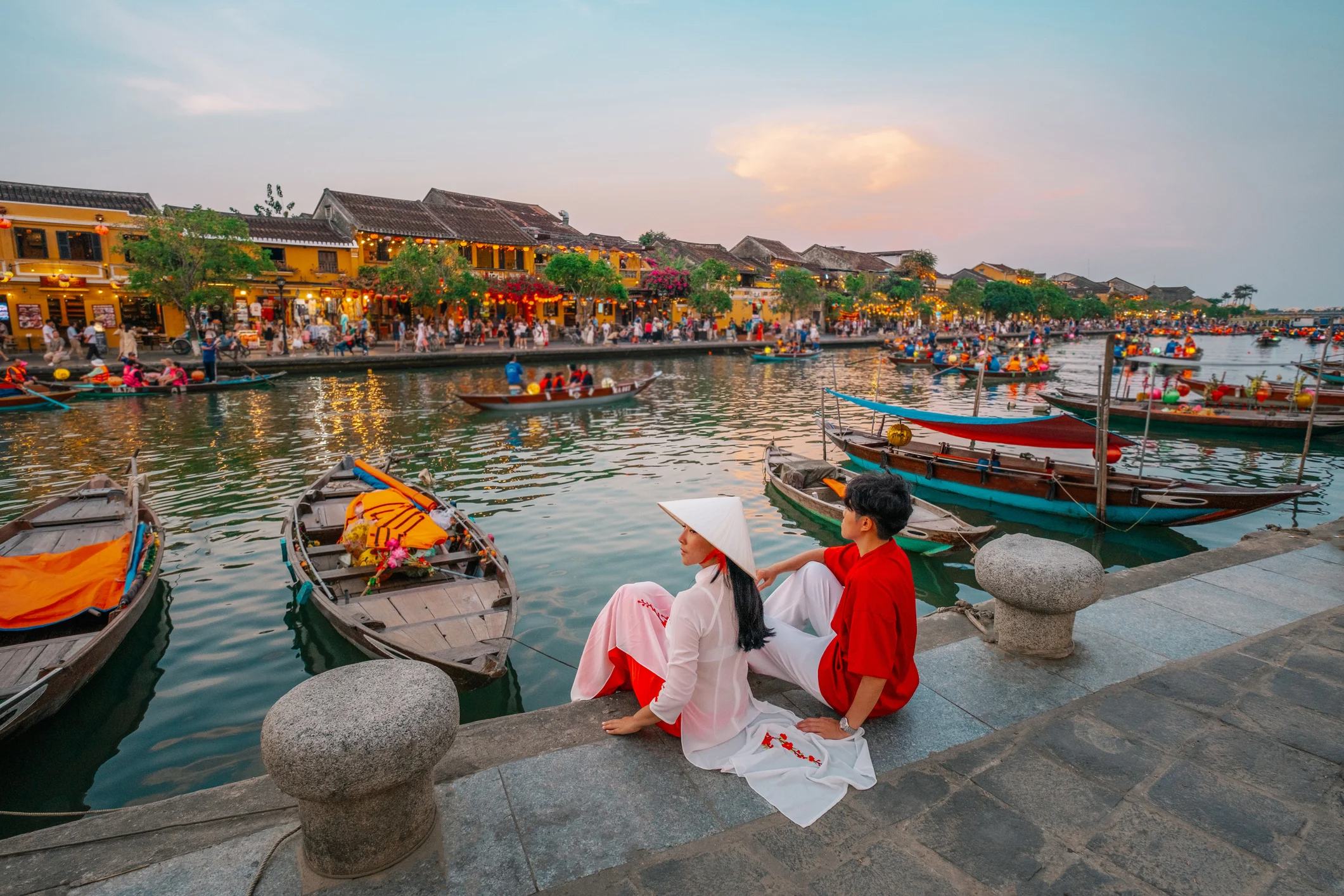 A couple sits by a riverbank in Hoi An, Vietnam, surrounded by boats and lively market scenes at sunset.