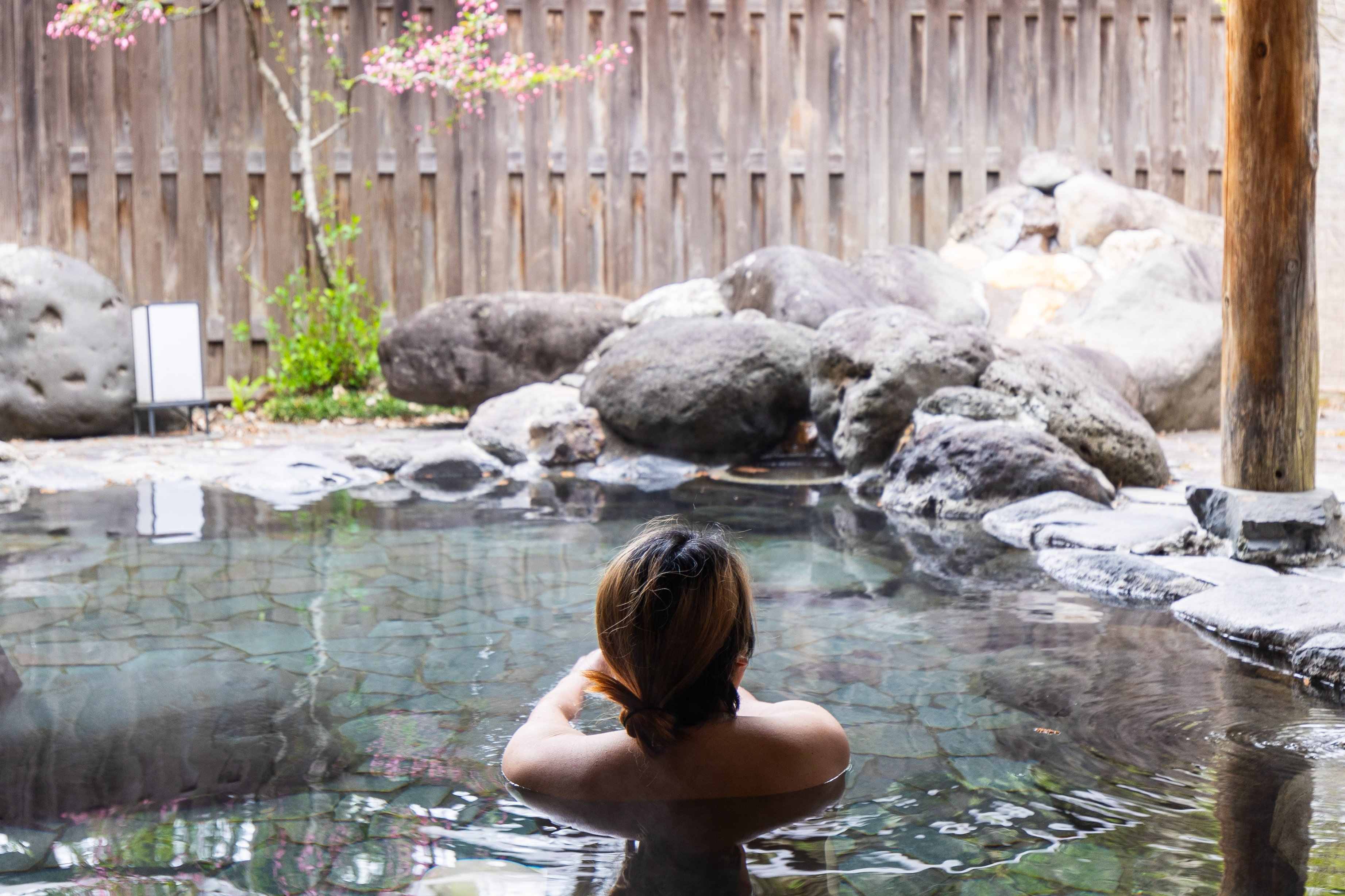 A person with shoulder-length hair relaxes in a stone hot spring surrounded by rocks and greenery.