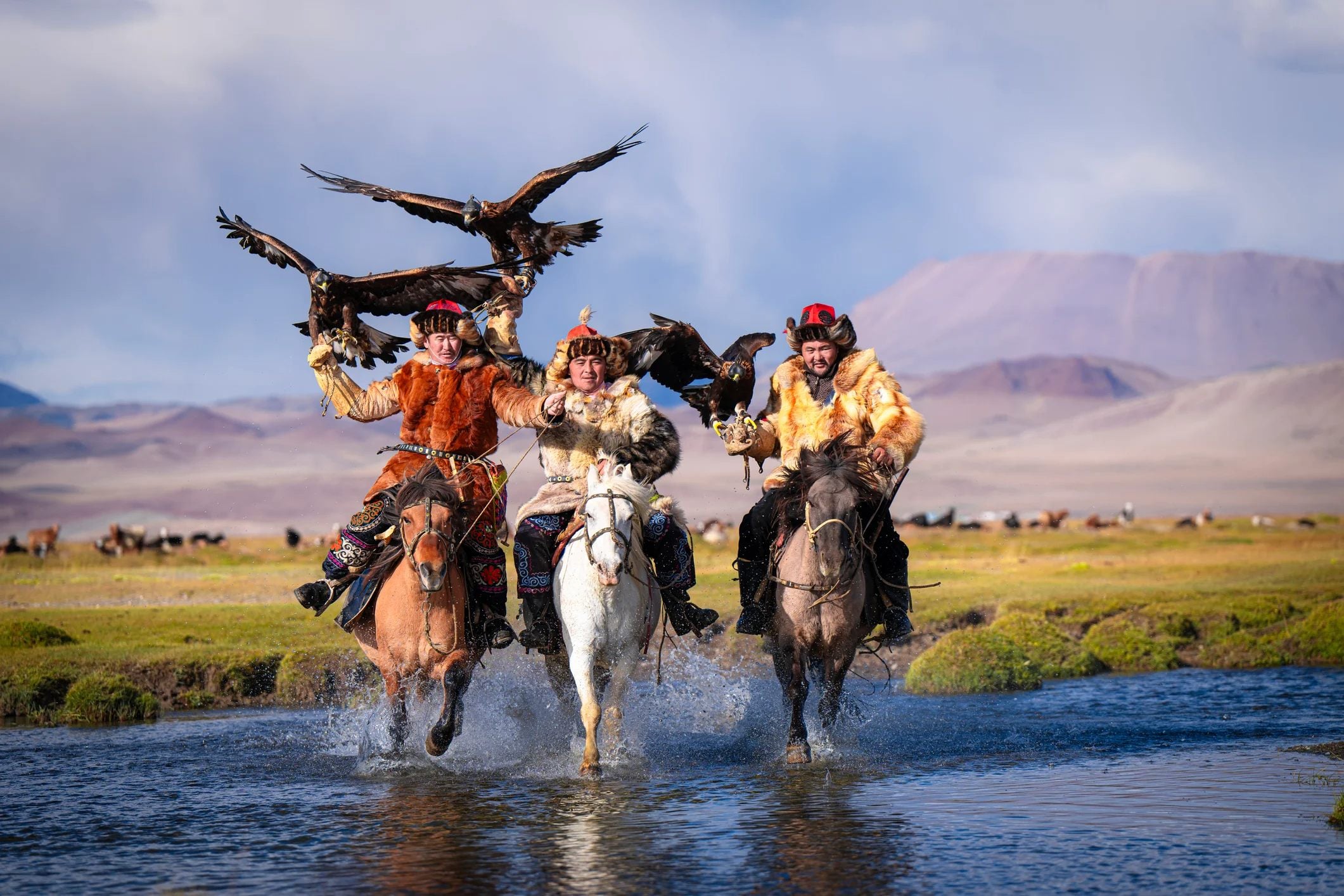 Three riders on horseback splash through a stream, each holding a golden eagle, with mountains in the background.