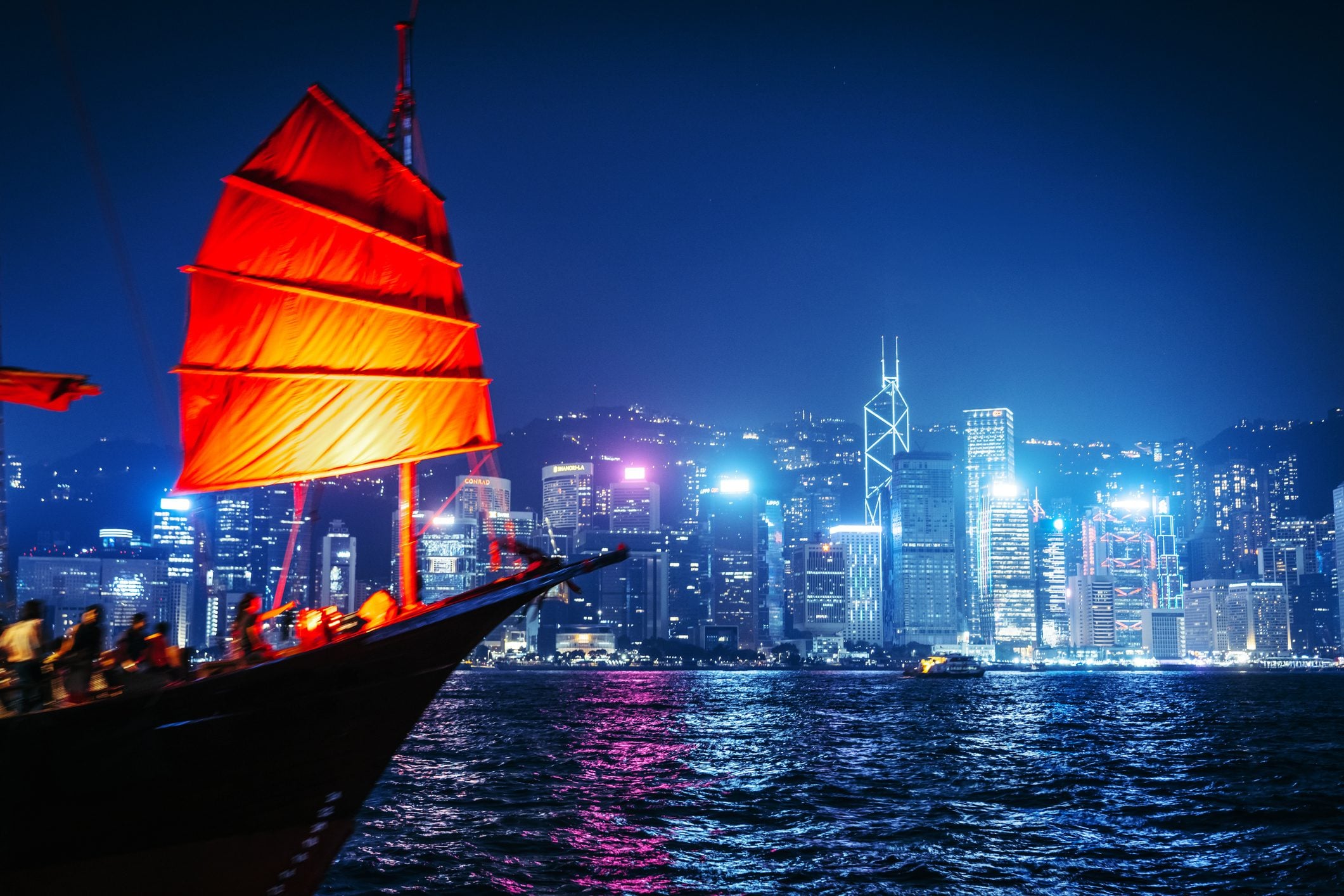 A traditional red-sailed boat in the foreground, with Hong Kong's illuminated skyline in the background at night.