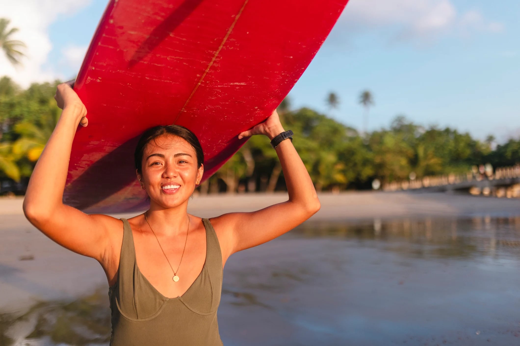 A smiling woman holds a red surfboard overhead at the beach, with palm trees and water in the background.
