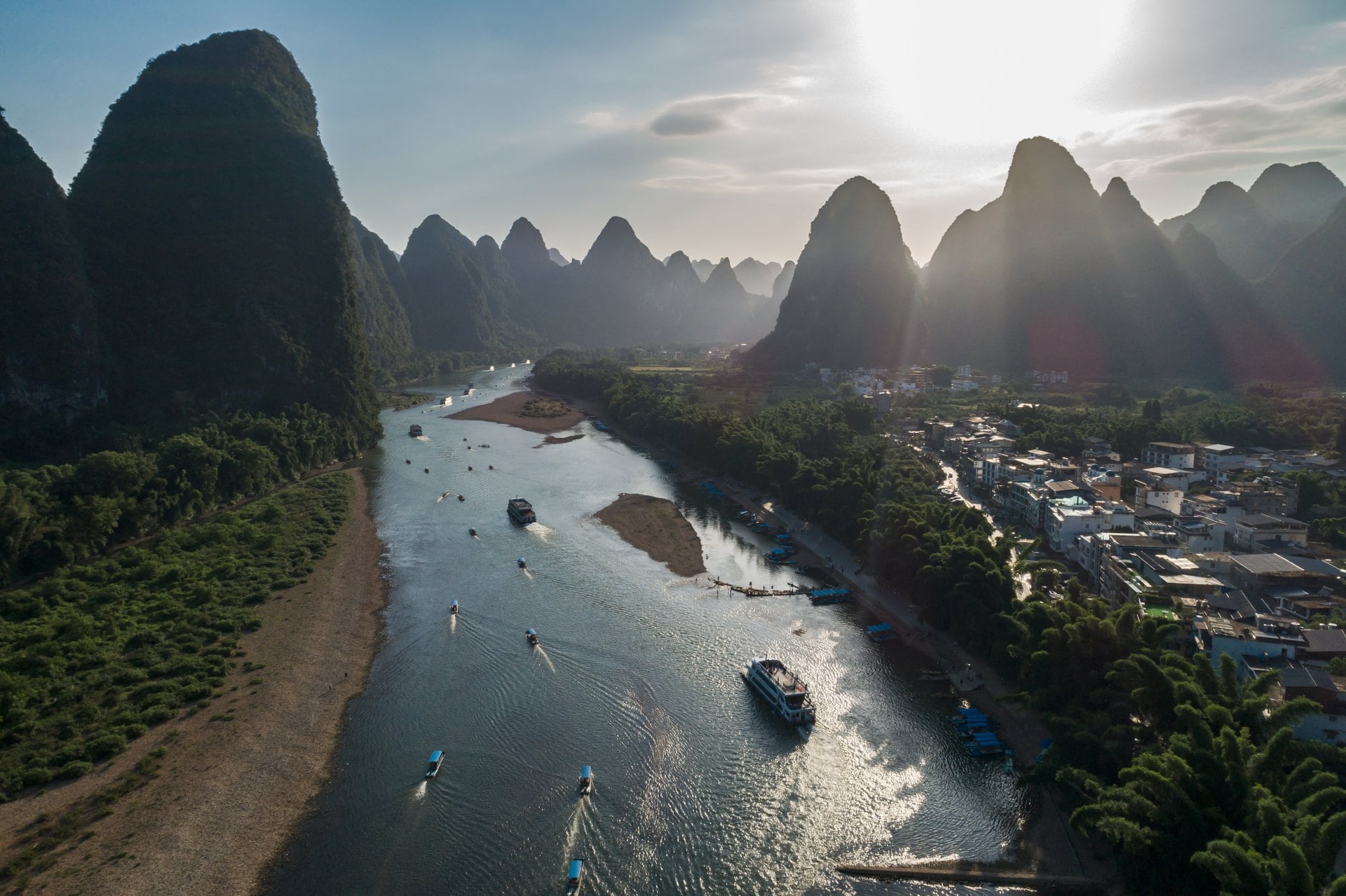 Aerial view of a river surrounded by lush mountains and a small town, with boats navigating the water in the foreground.
