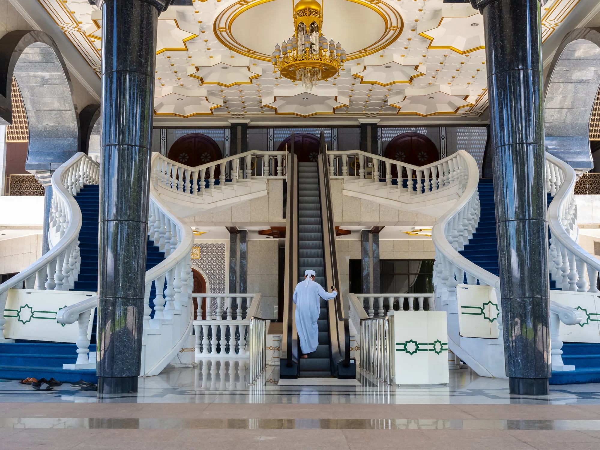 A person in traditional attire stands on an escalator in an ornate hall with grand staircases and a beautifully decorated ceiling.