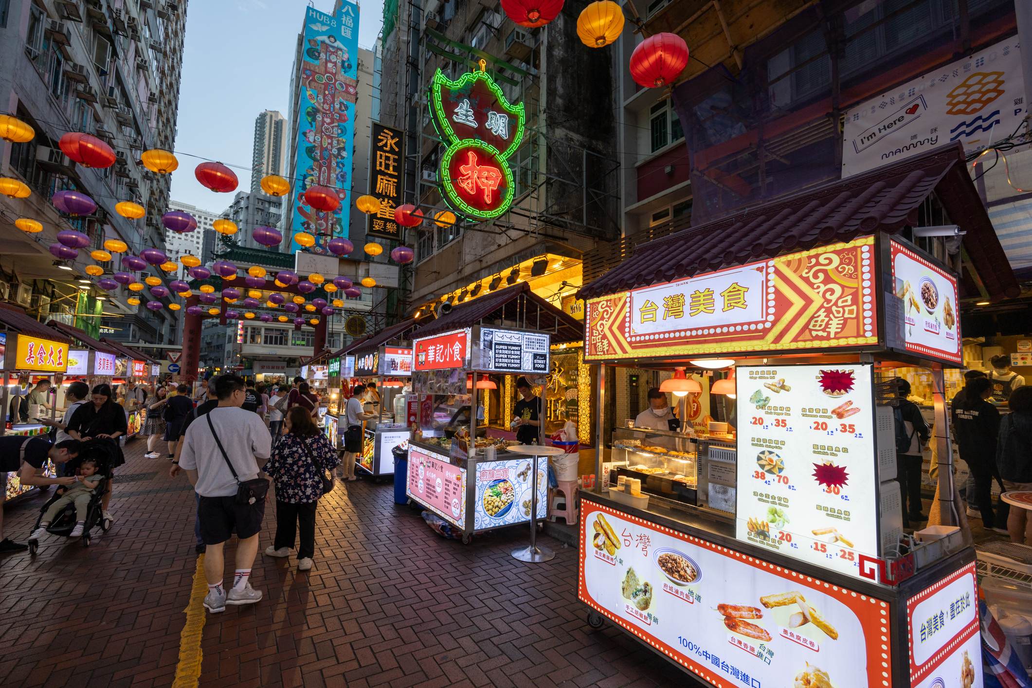 Vibrant street market scene with food stalls, colorful lanterns overhead, and people walking among the stalls.