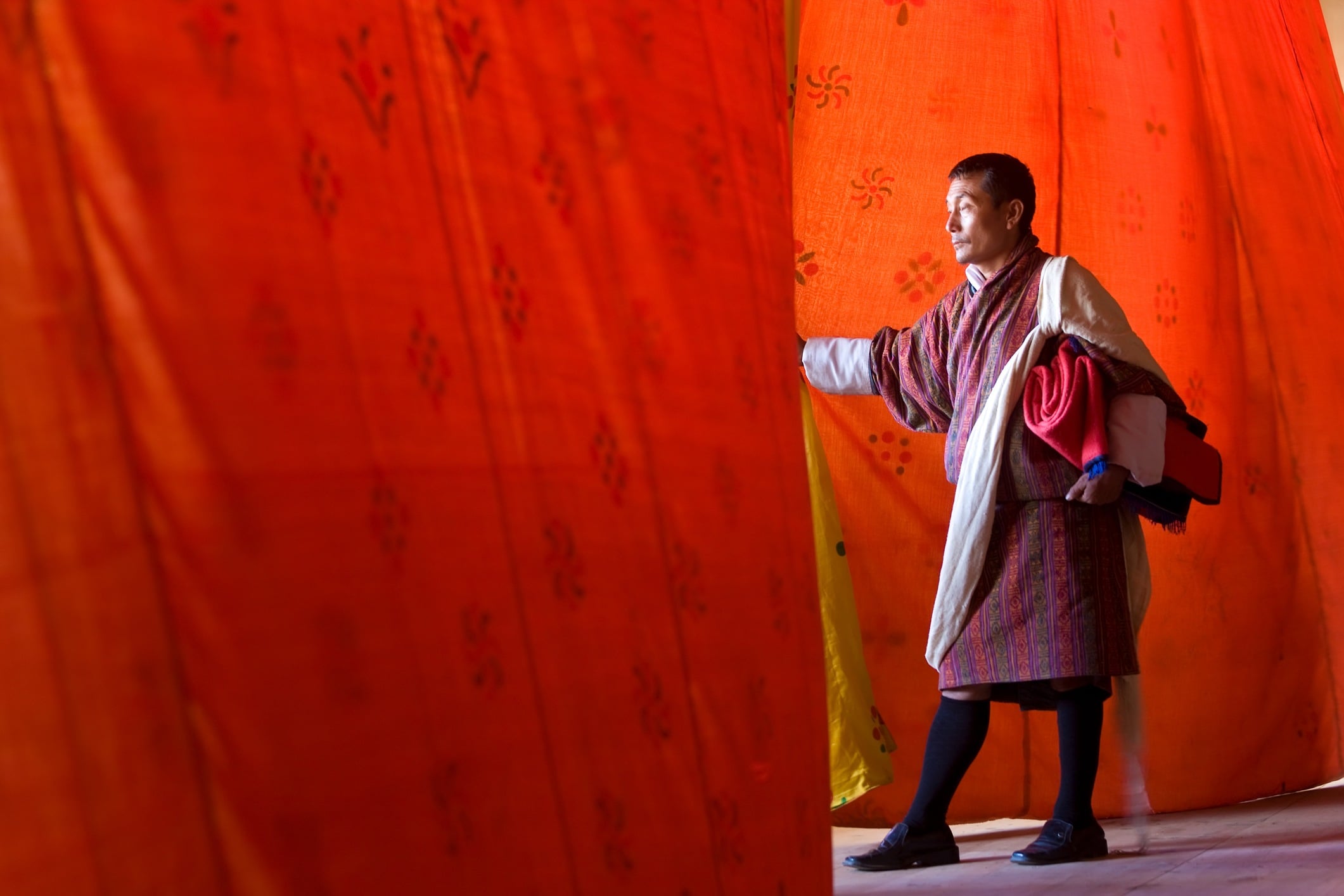 A man in traditional attire stands near colorful fabric, reaching out as he appears to enter a bright space.