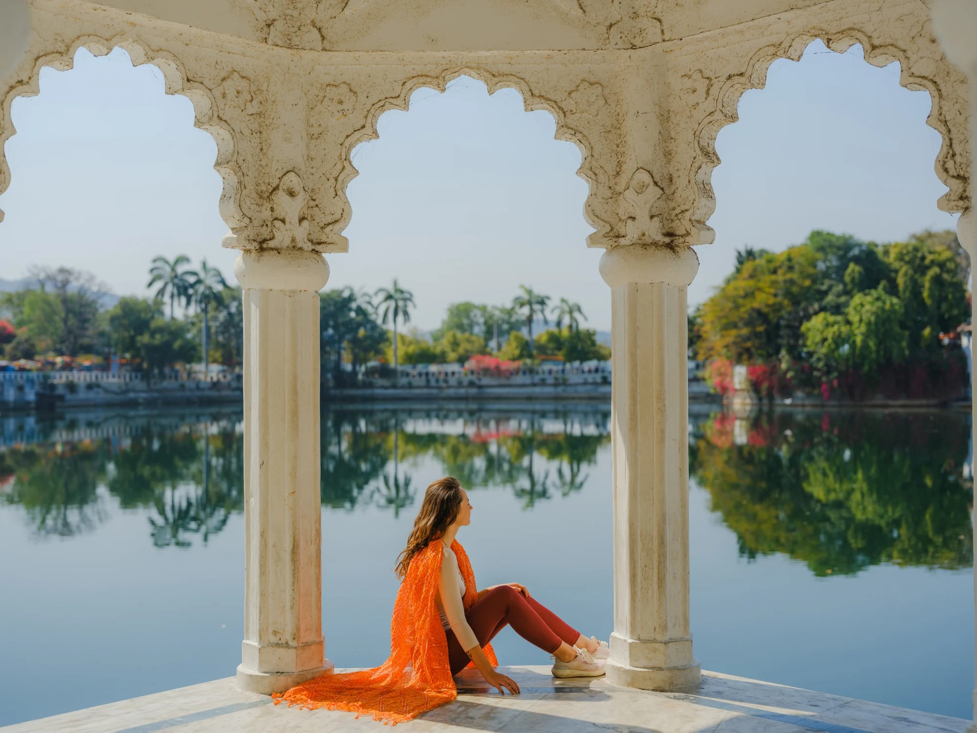 A woman in an orange outfit sits by a serene lake, framed by ornate white pillars and lush greenery in the background.