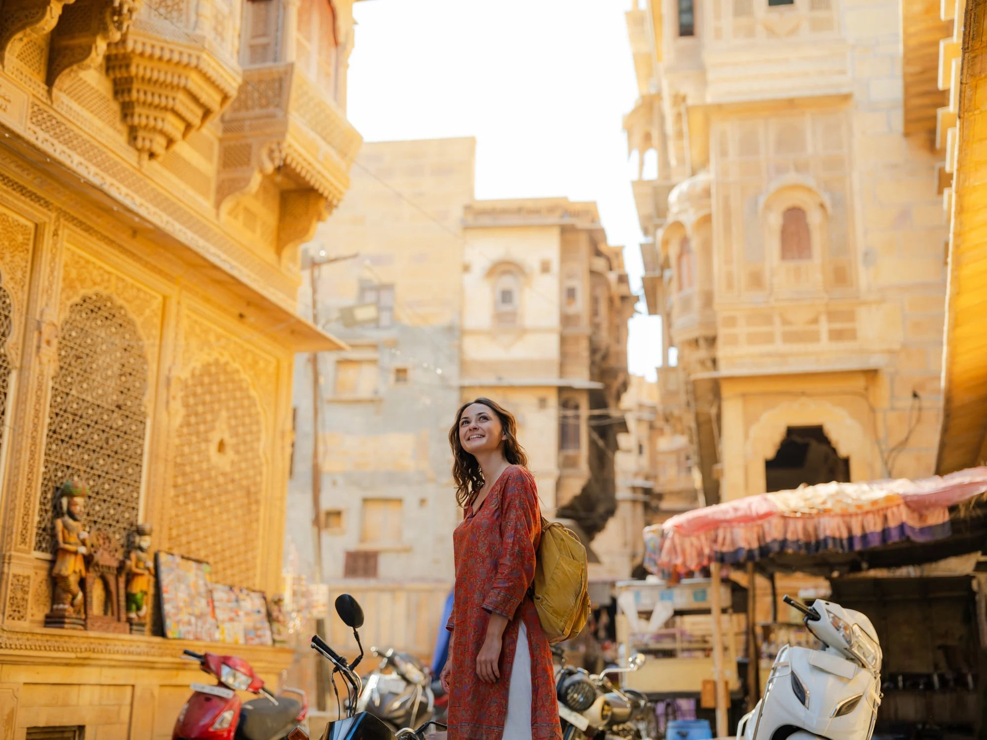 A woman in an orange dress stands in a sunlit narrow street, surrounded by historic buildings and parked motorcycles.