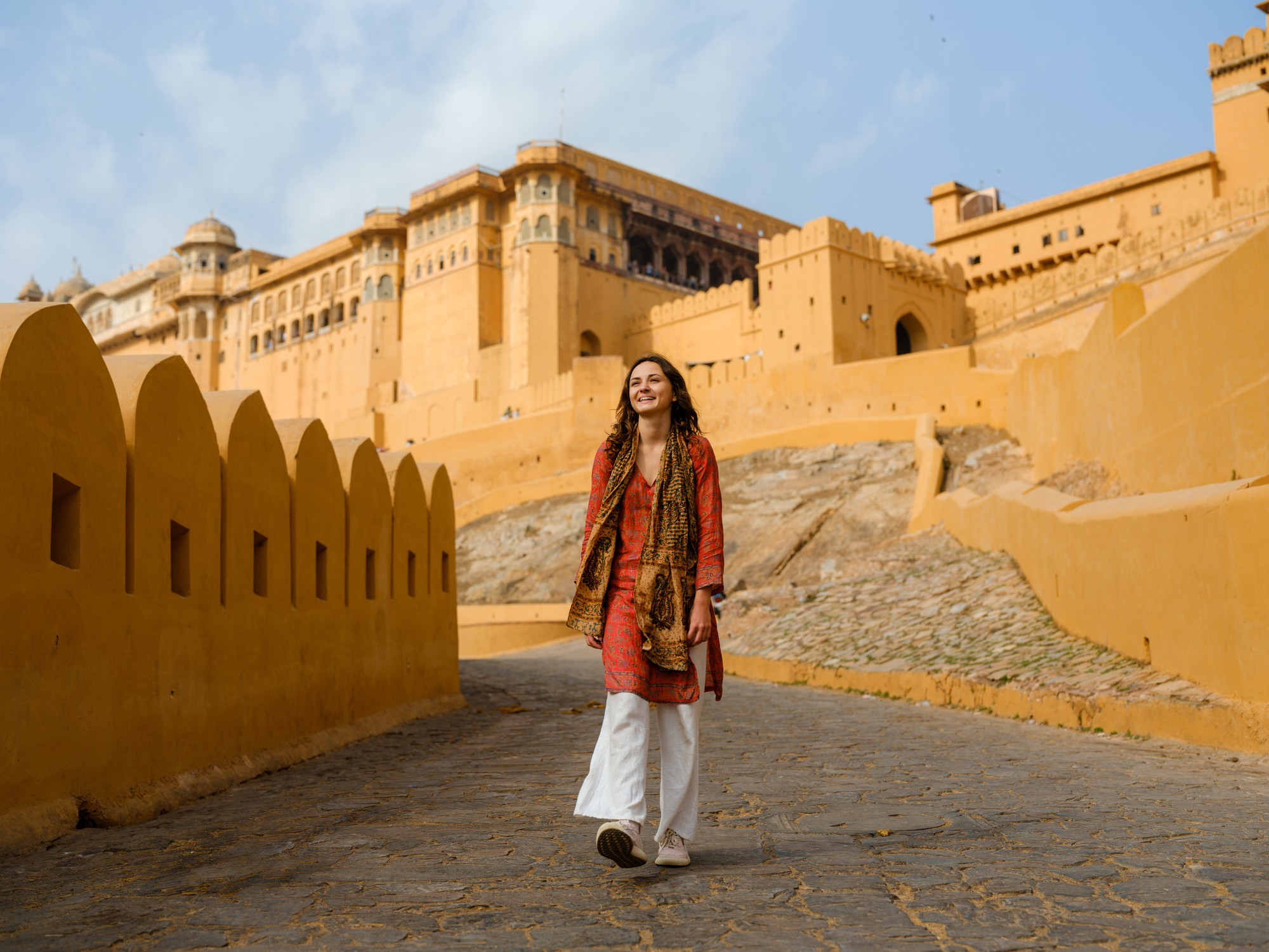 A woman walks along a stone path toward a large, orange fort against a blue sky.