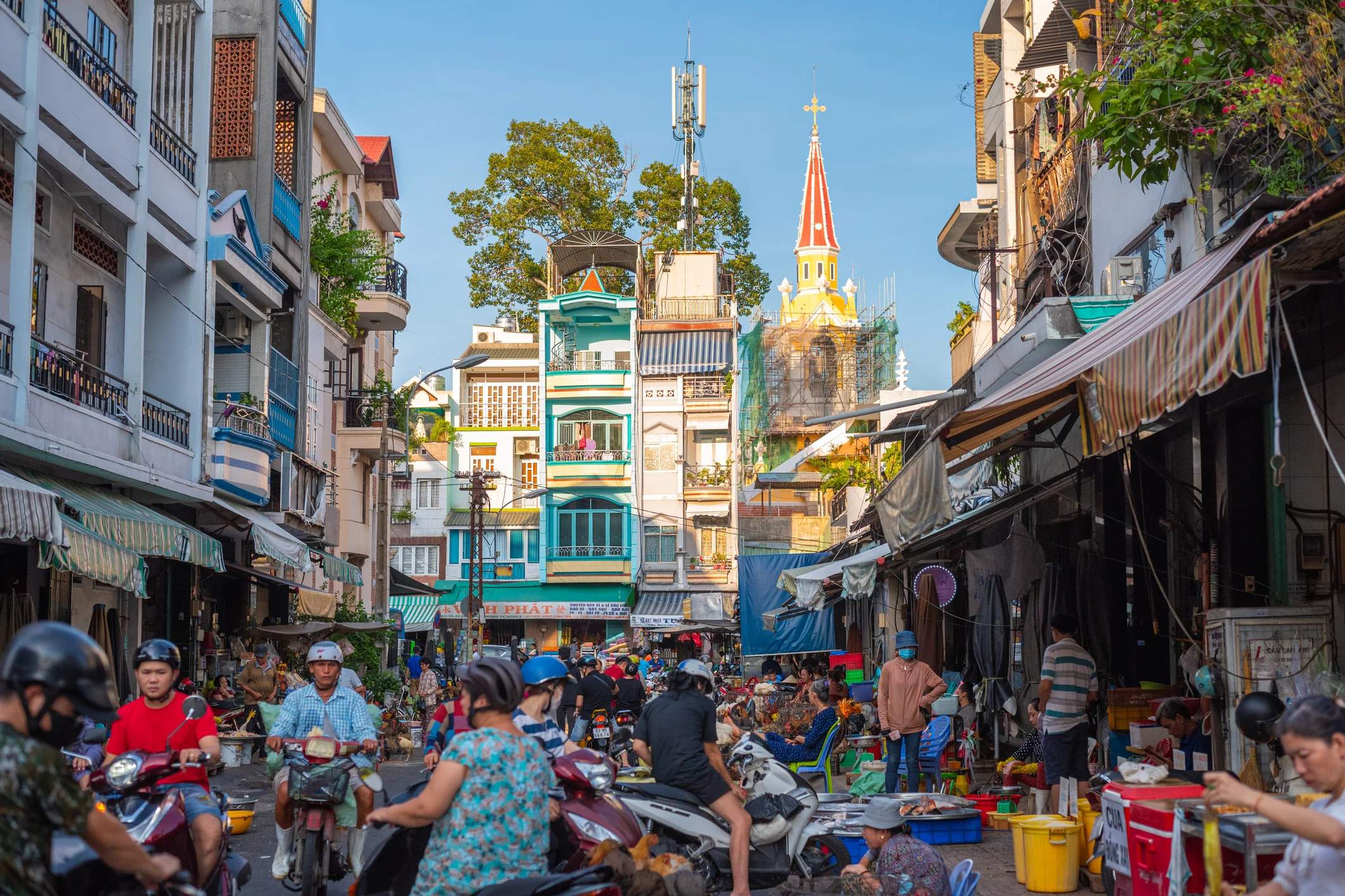 Bustling street scene filled with people and motorbikes, tall buildings, and a church steeple in the background.