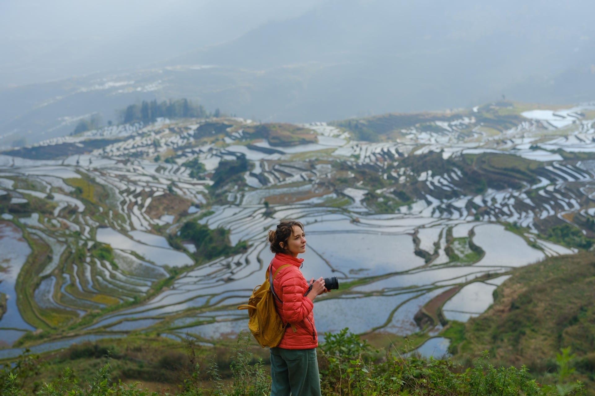 Person in a red jacket stands on a hillside, overlooking terraced rice fields reflecting water under a cloudy sky.