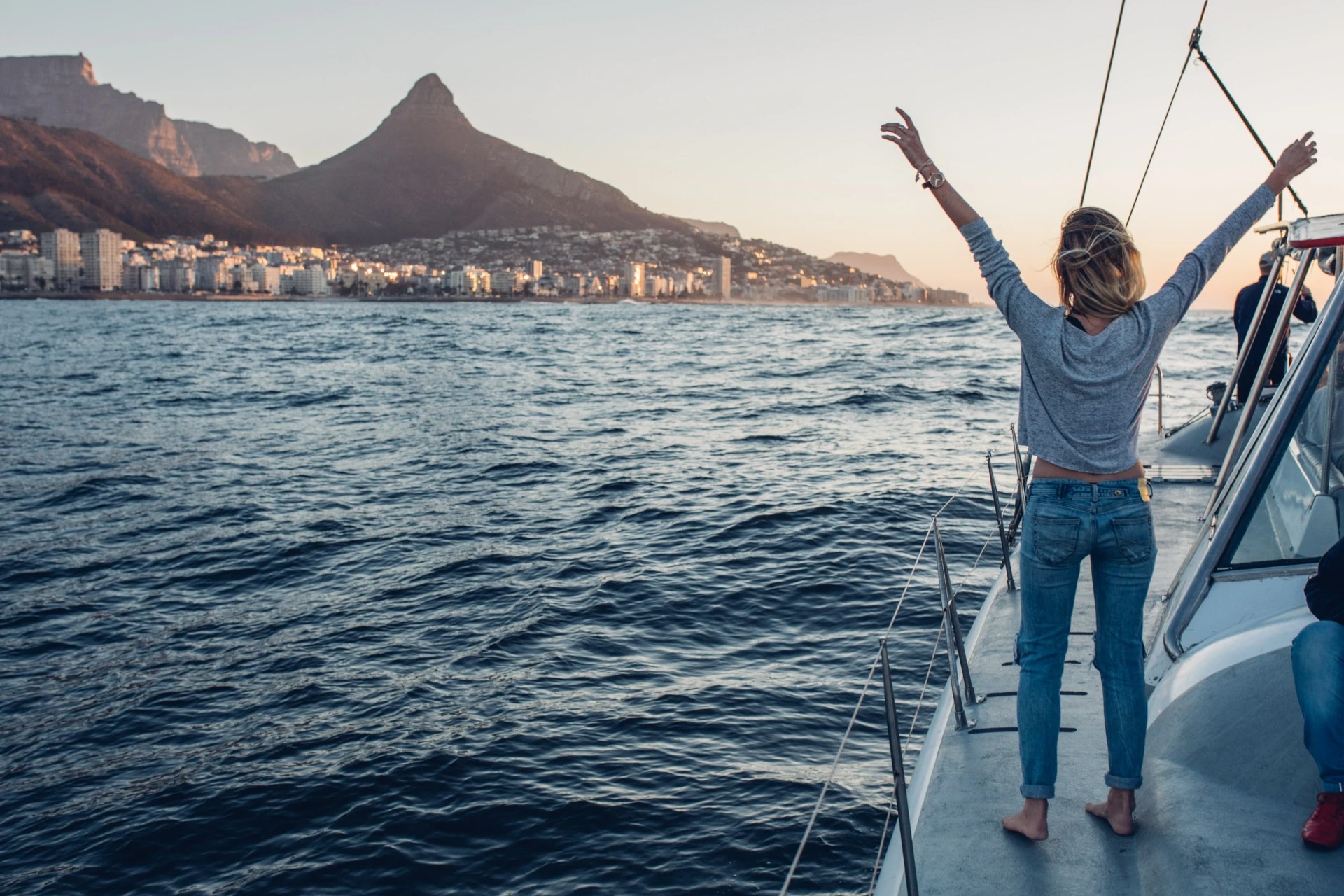 Person on a sailboat raises arms in joy, with mountains and a city skyline in the background at sunset.