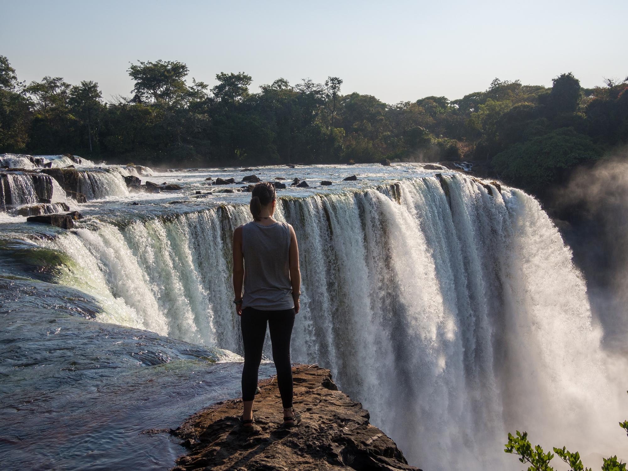 A person stands on the edge of a rocky cliff, looking out at a large waterfall surrounded by lush greenery.