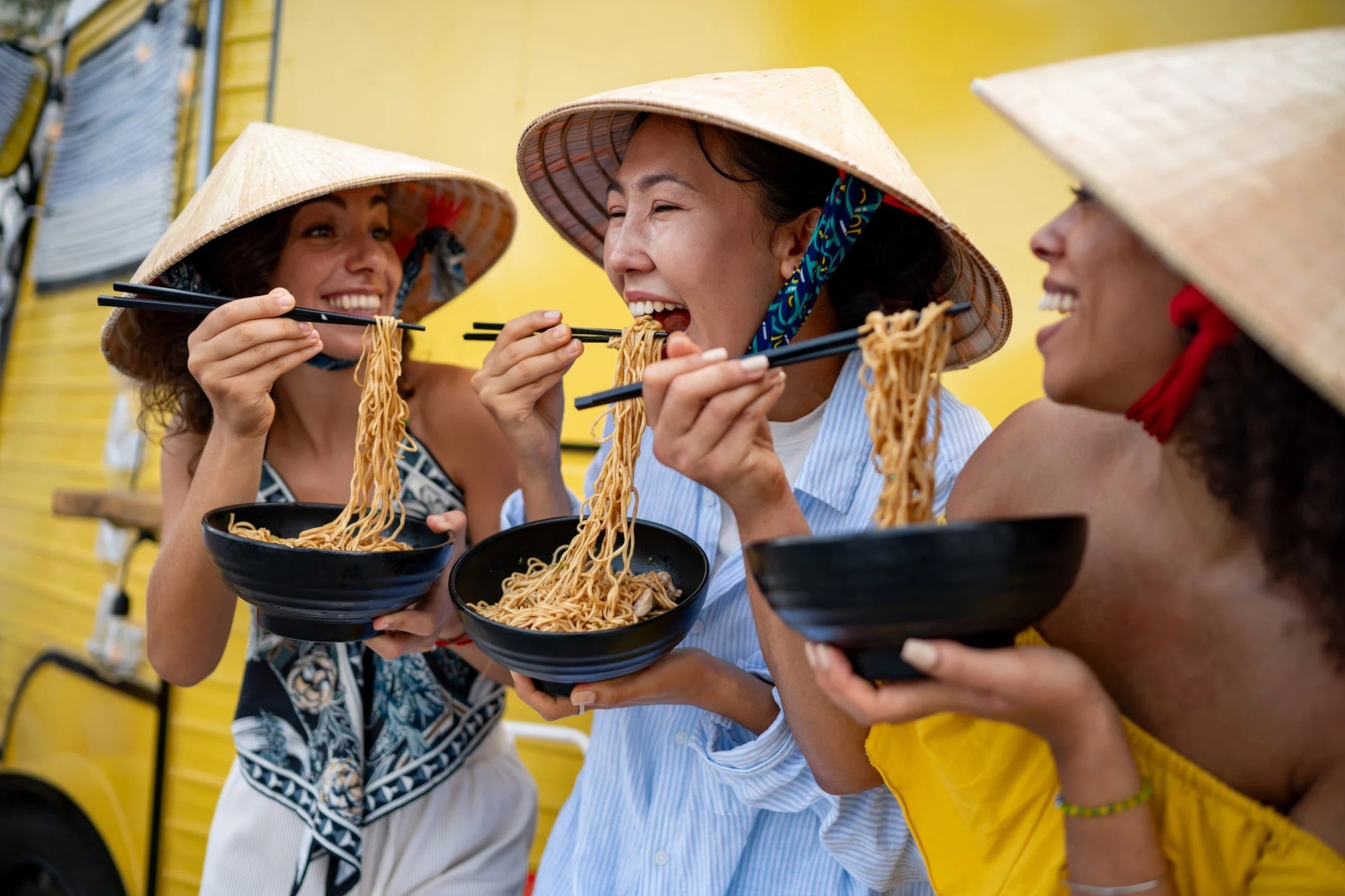 Three women in traditional hats enjoy noodles from bowls, laughing together in front of a bright yellow food truck.