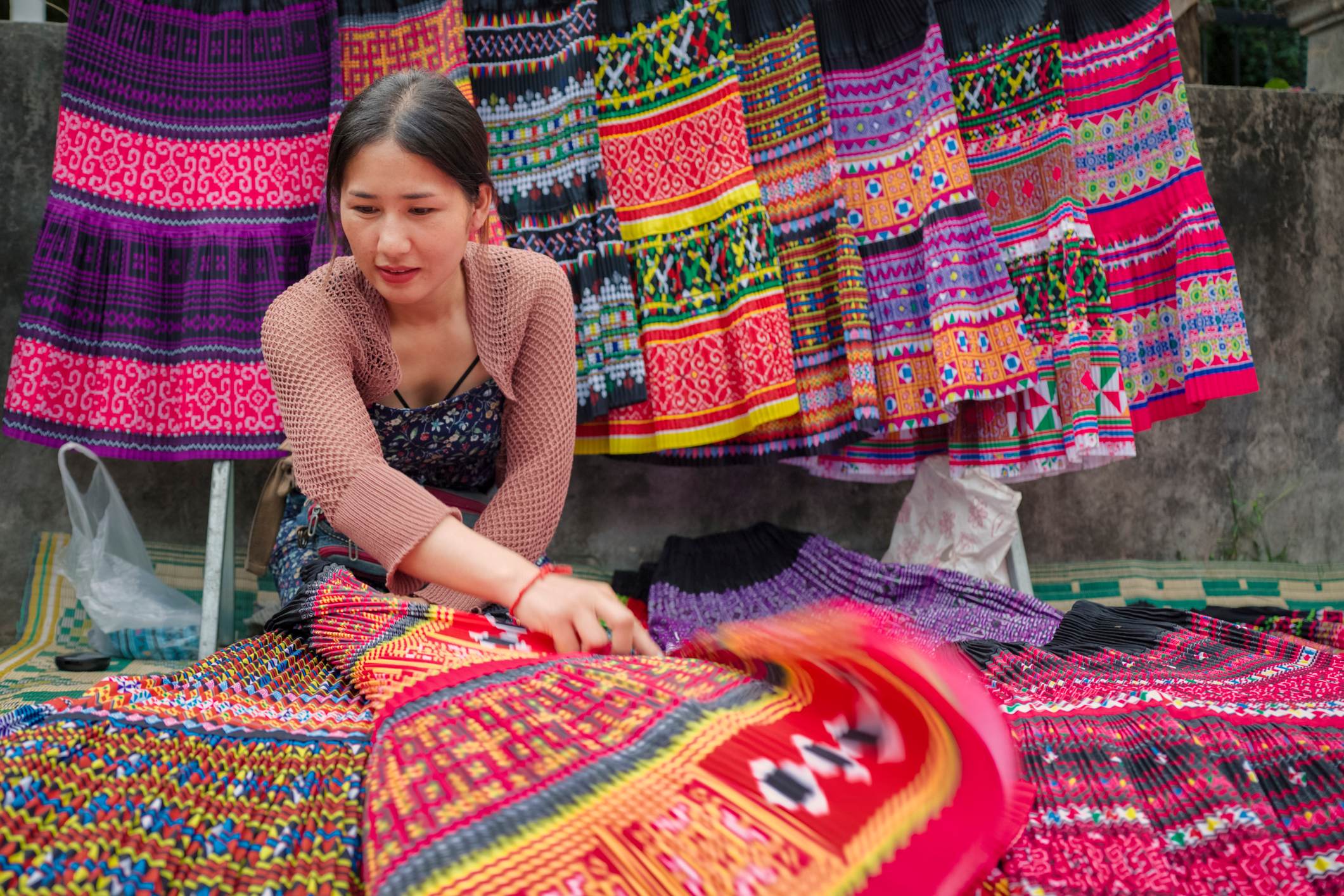A woman arranges colorful, patterned textiles at a market, with vibrant traditional garments hanging in the background.