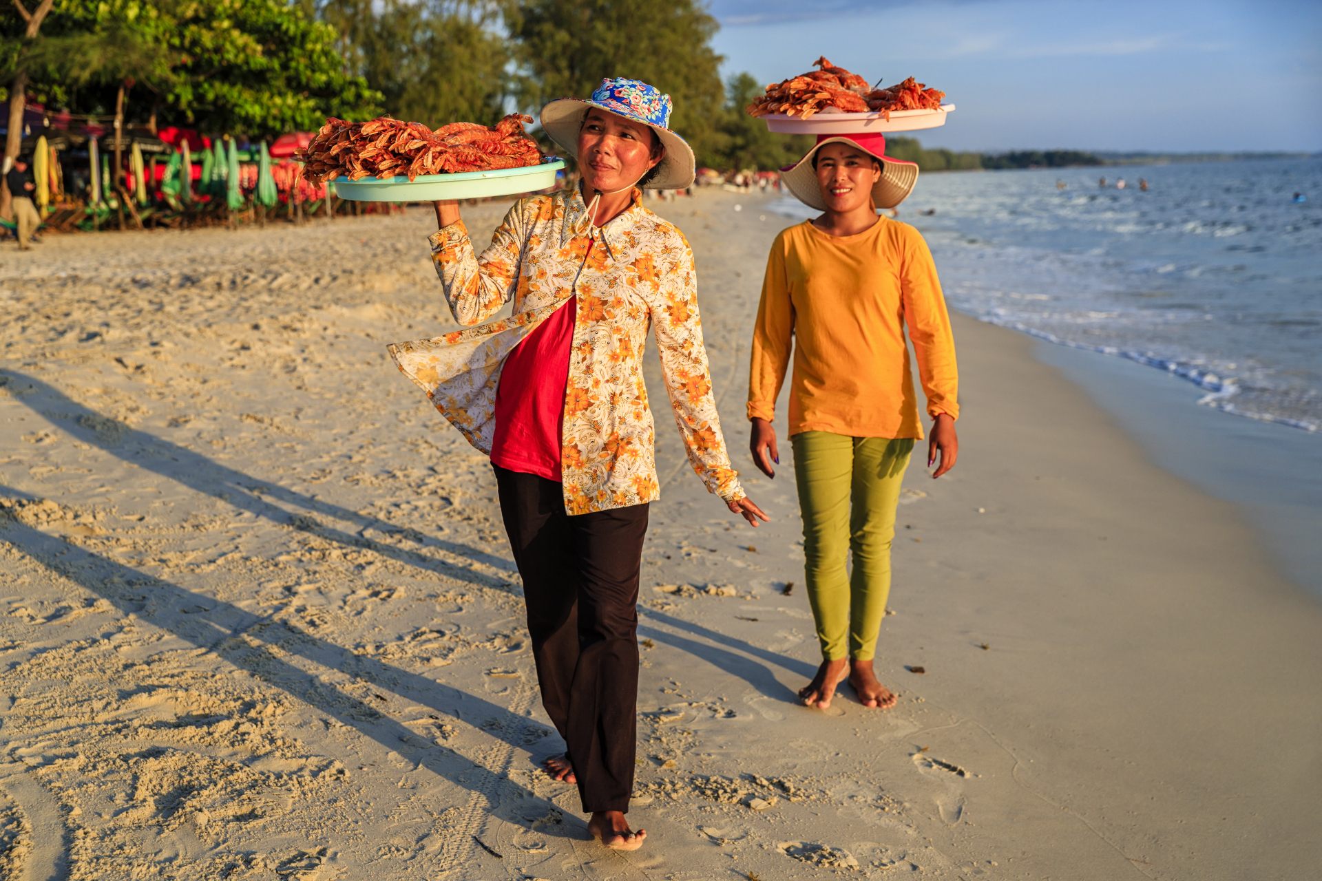 Two women walk along a beach, carrying trays of food on their heads, with a calm sea and trees in the background.