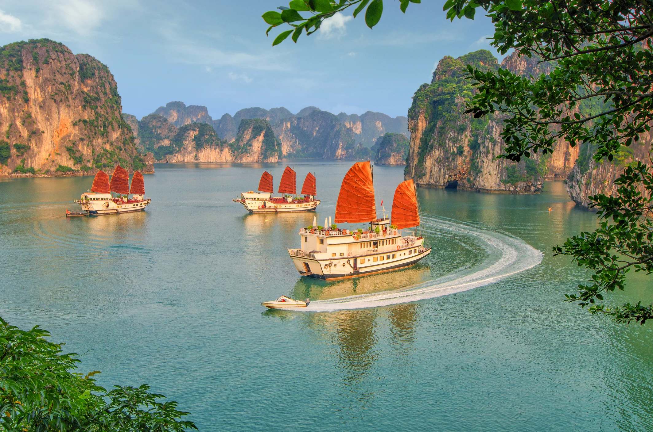 Three traditional wooden boats with red sails glide through a serene bay surrounded by lush green cliffs.