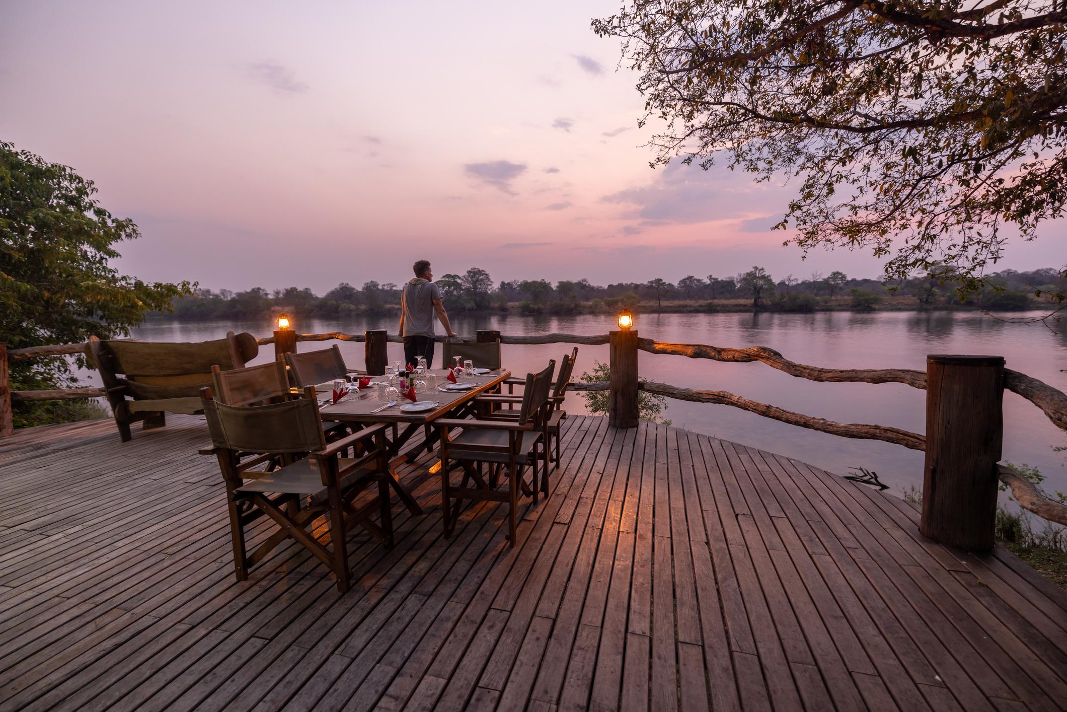 A wooden deck by a river at sunset, with a dining table set and a person standing in the background.