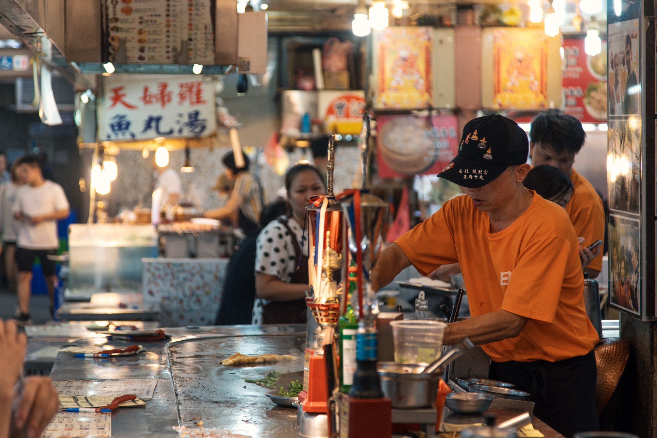 A busy food stall with people, a vendor in an orange shirt cooking, and a woman in polka dots at a table.