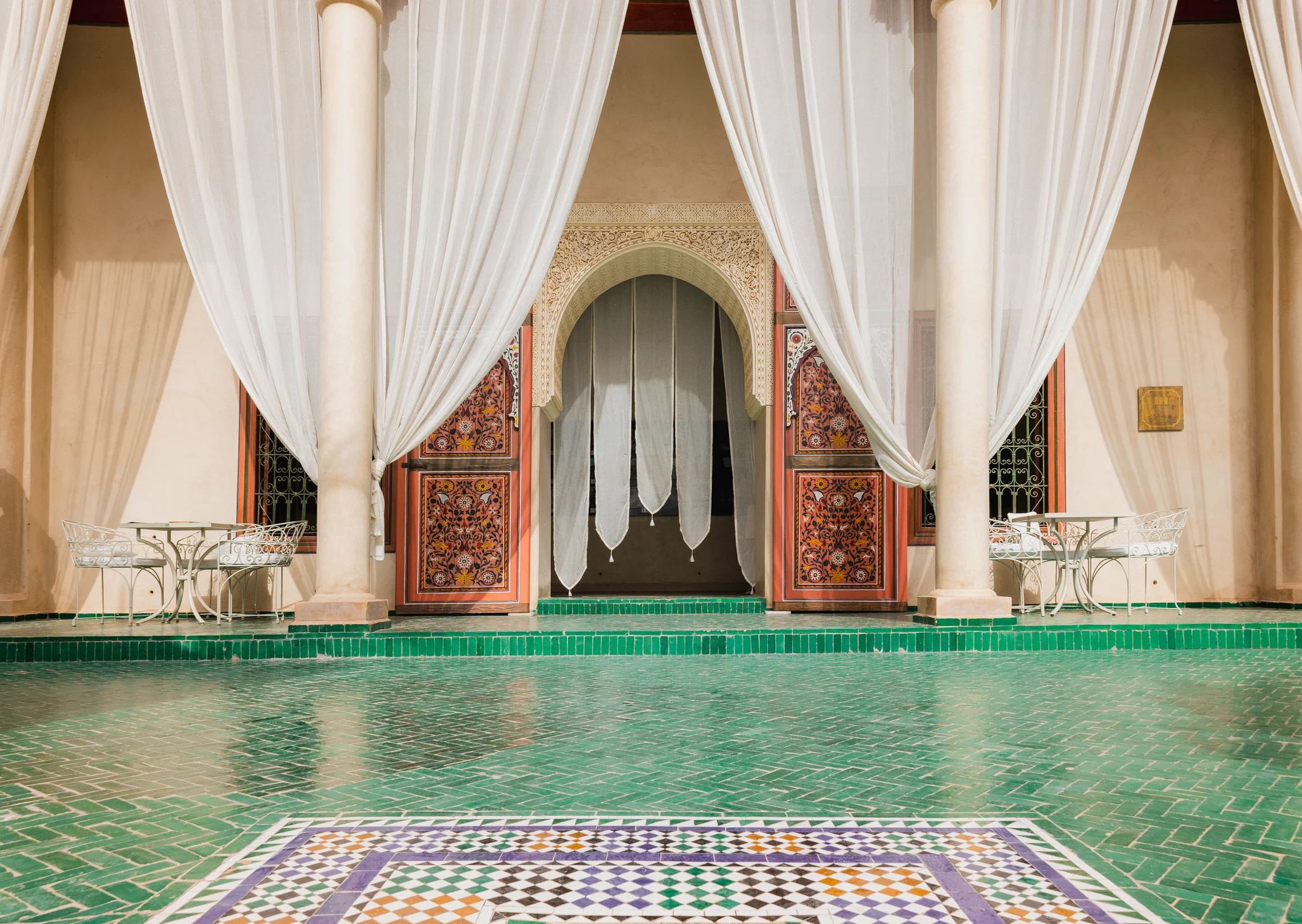 Elegant interior with green tiled floor, sheer drapes, and intricately carved wooden doors in a Moroccan-style setting.