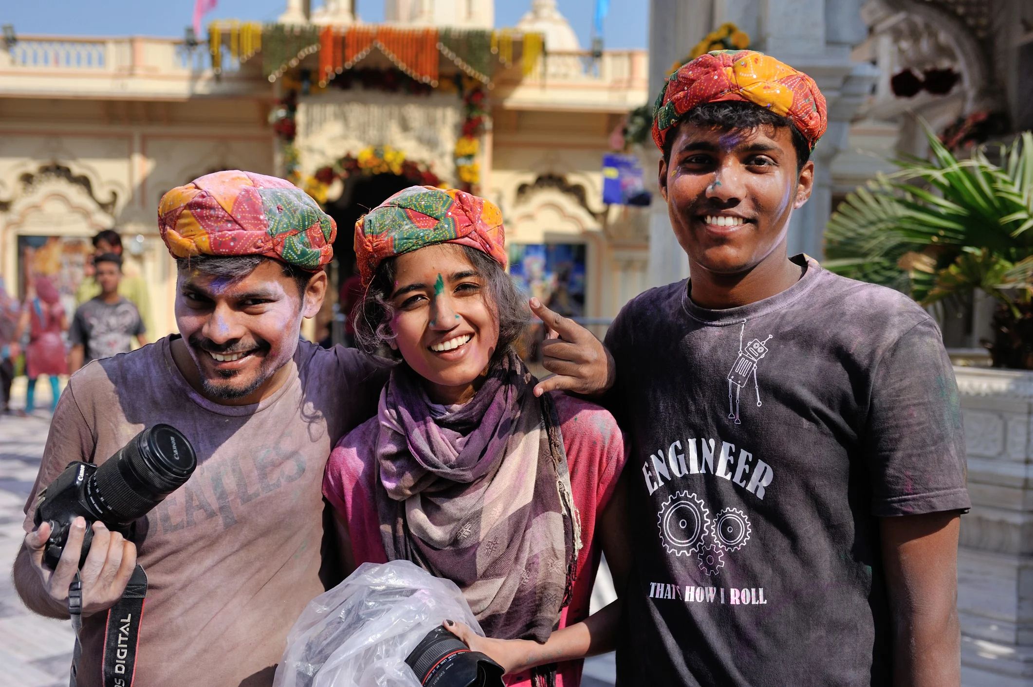 Three smiling friends in colorful turbans, holding cameras, pose together in a festive outdoor setting.