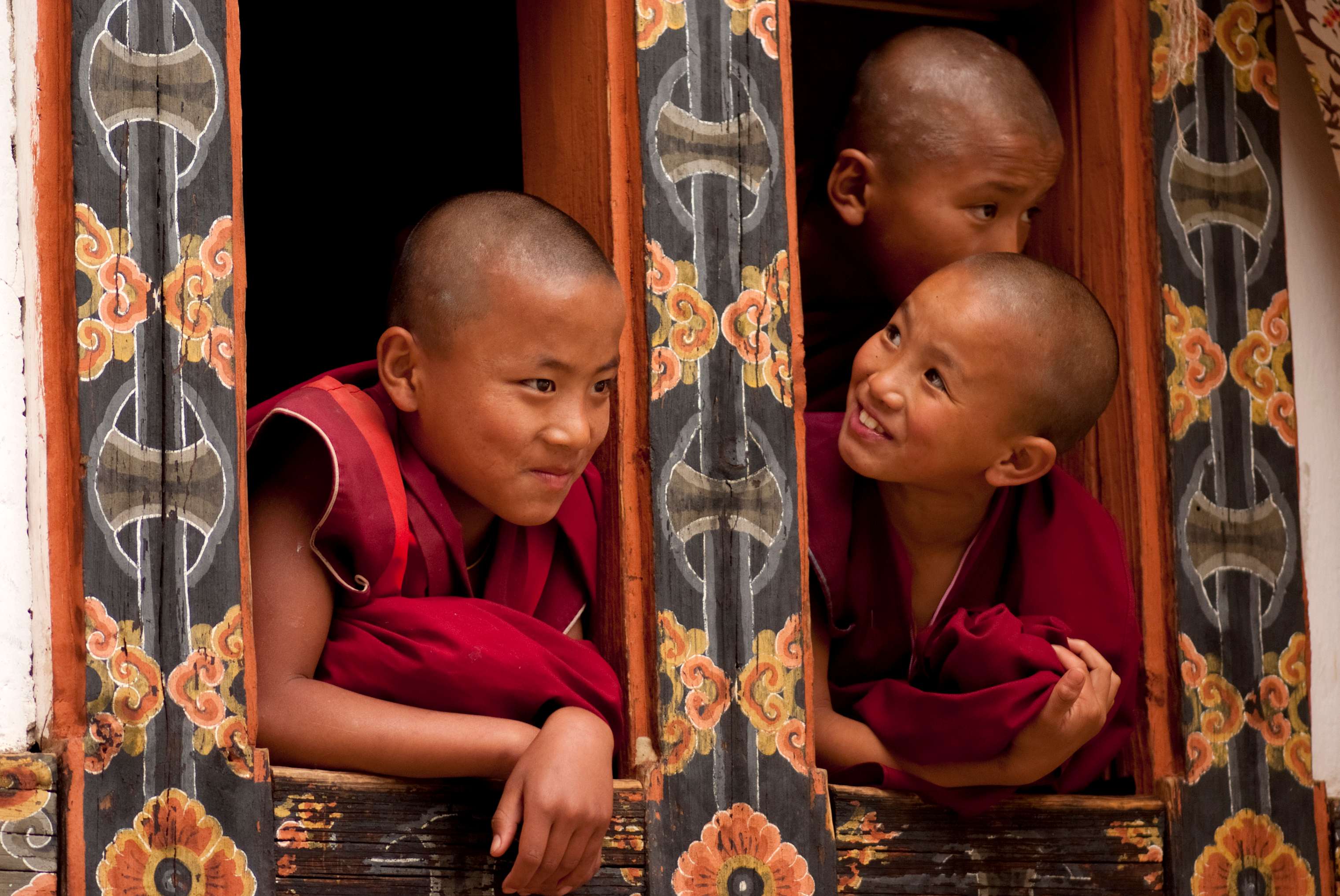 Three young monks with shaved heads peek out from a beautifully decorated window, smiling and engaging with each other.