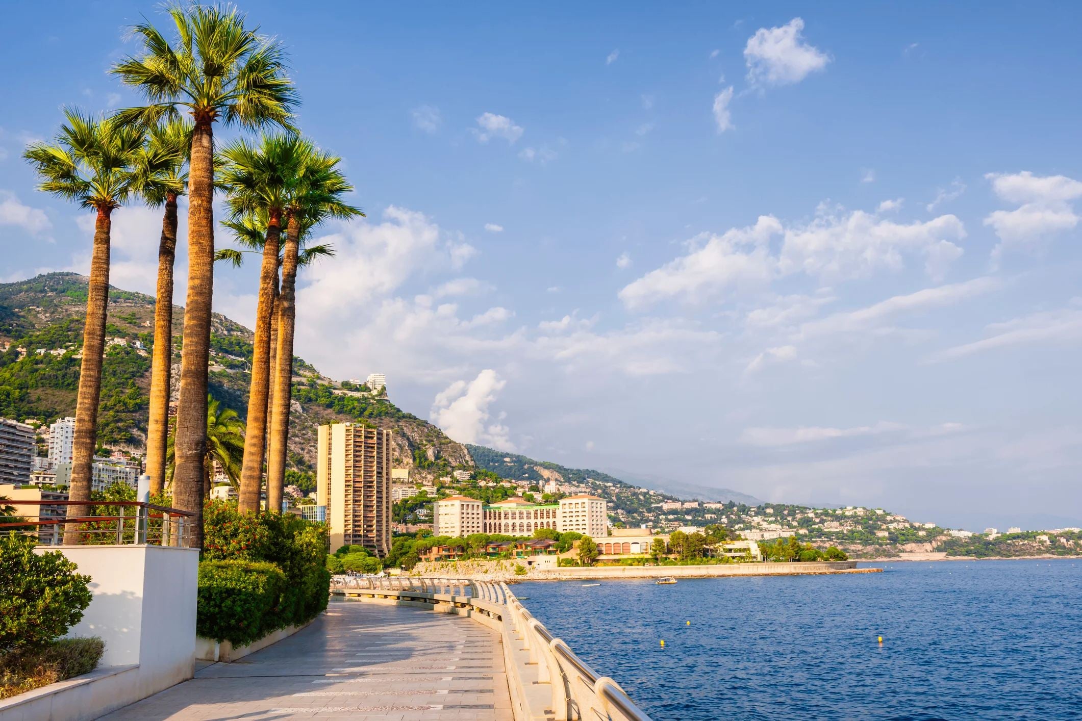 Scenic view of a coastal promenade lined with palm trees, buildings, and mountains under a blue sky with clouds.