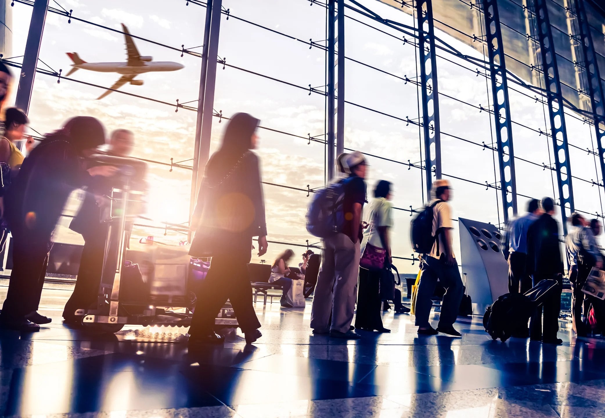 Blurred travelers moving through a bustling airport terminal, with an airplane flying outside the large glass windows.