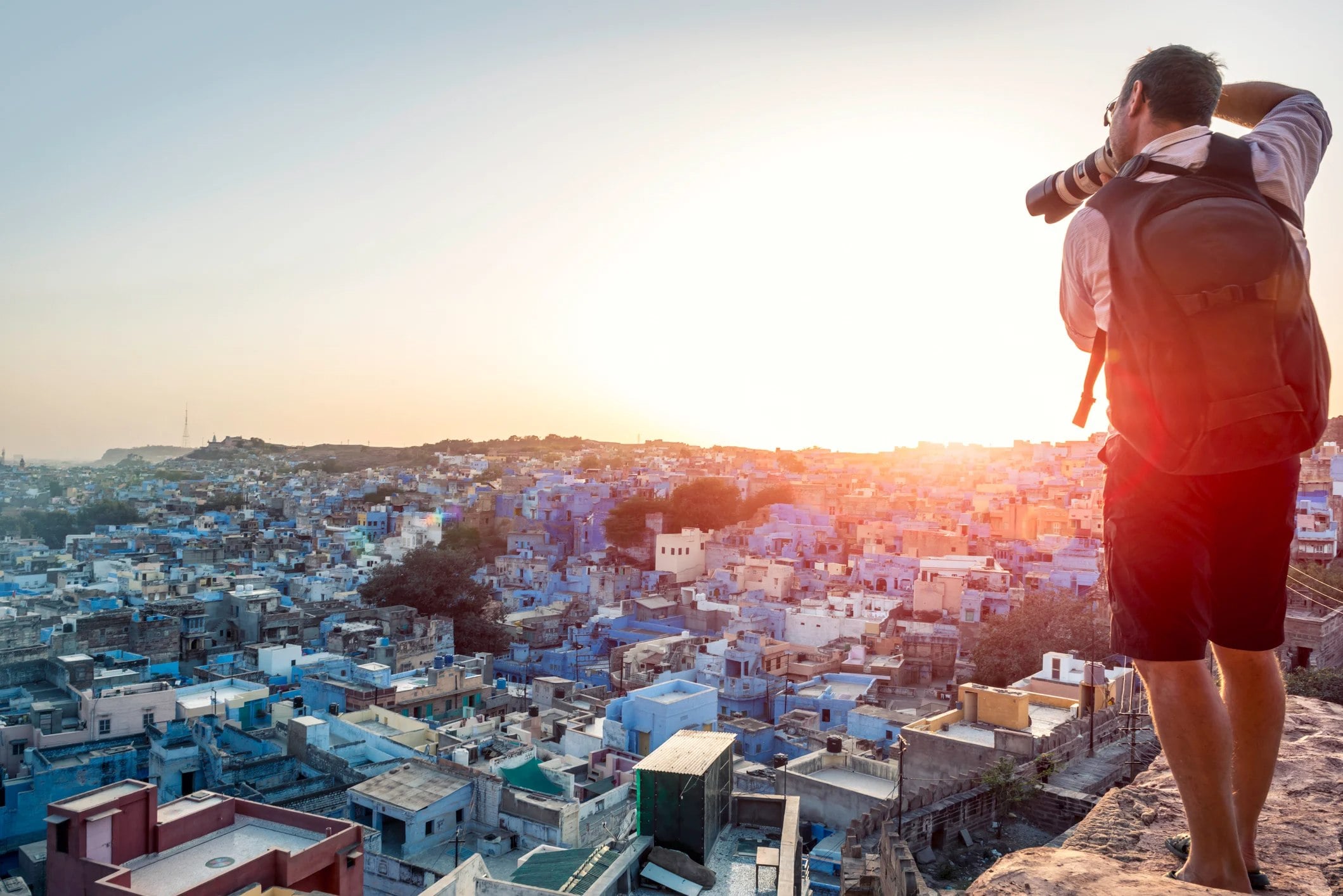 A photographer stands on a hilltop, capturing a sunset over a cityscape of blue buildings.