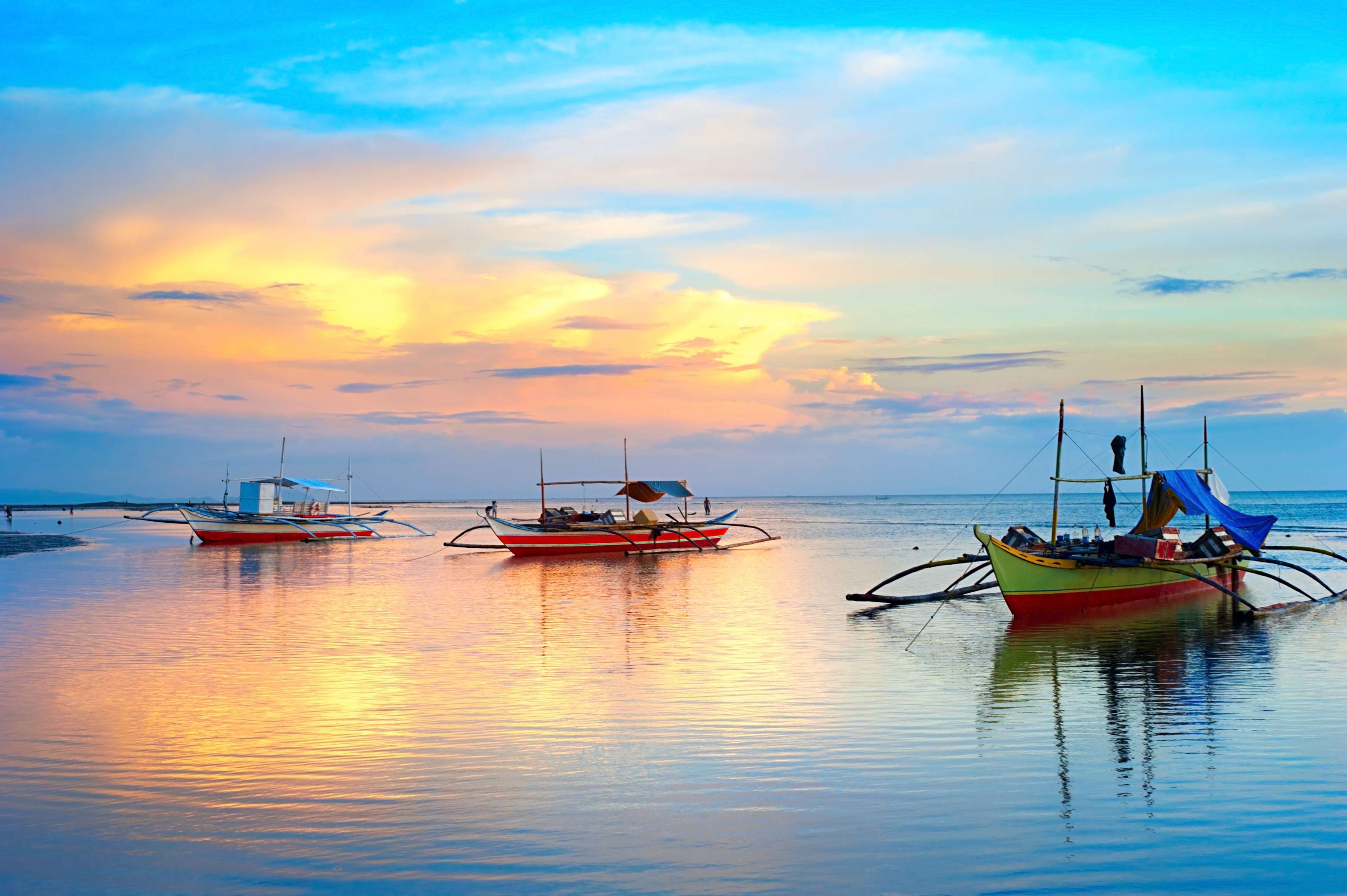 Three colorful boats on calm water under a vibrant sunset sky with clouds reflecting in the water.