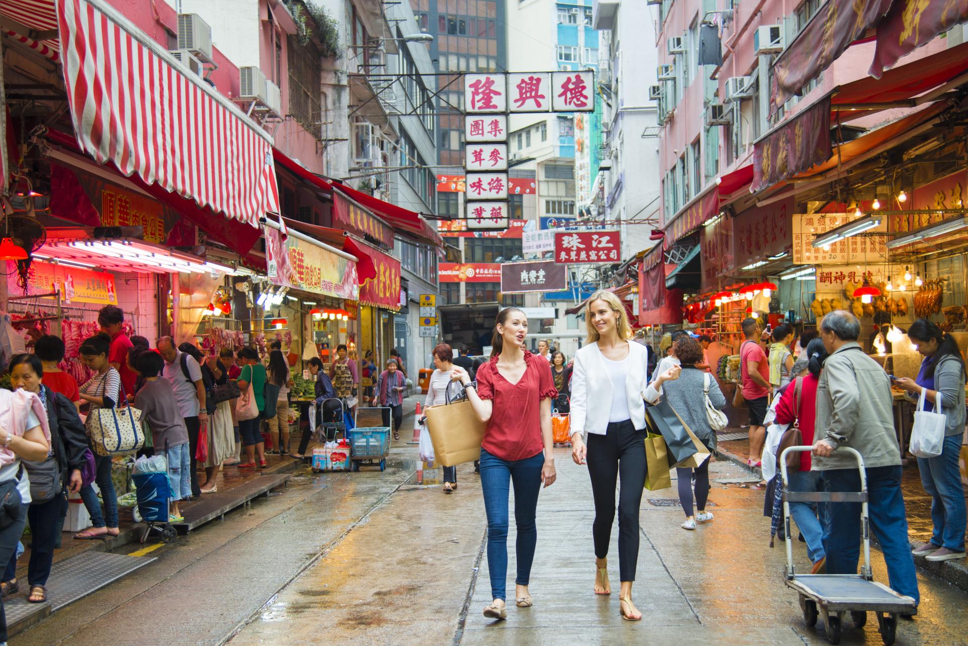 Two women walk through a bustling market street filled with colorful shops and people, vibrant signs in the background.