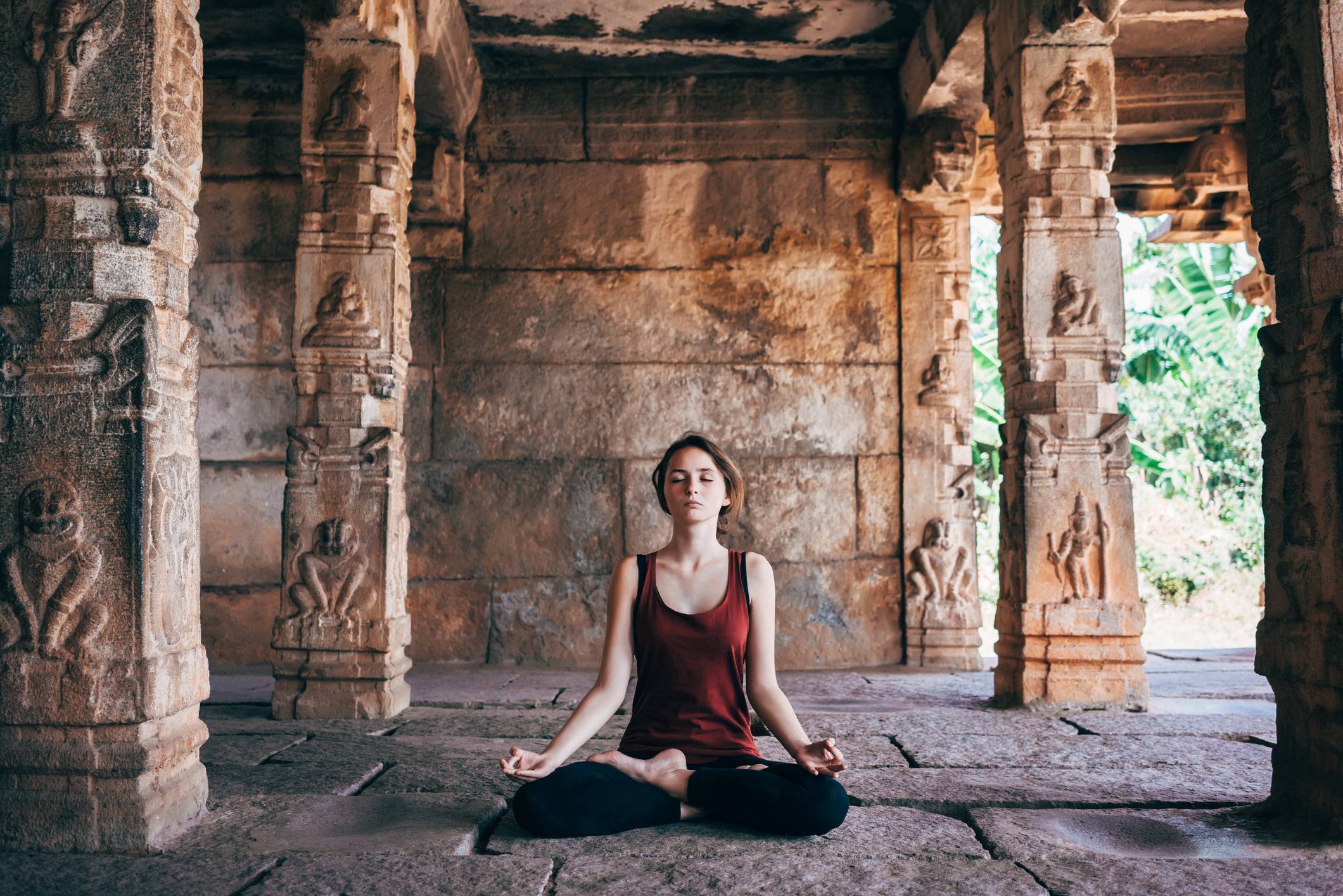 A woman meditating in a serene temple with stone pillars and lush greenery in the background.