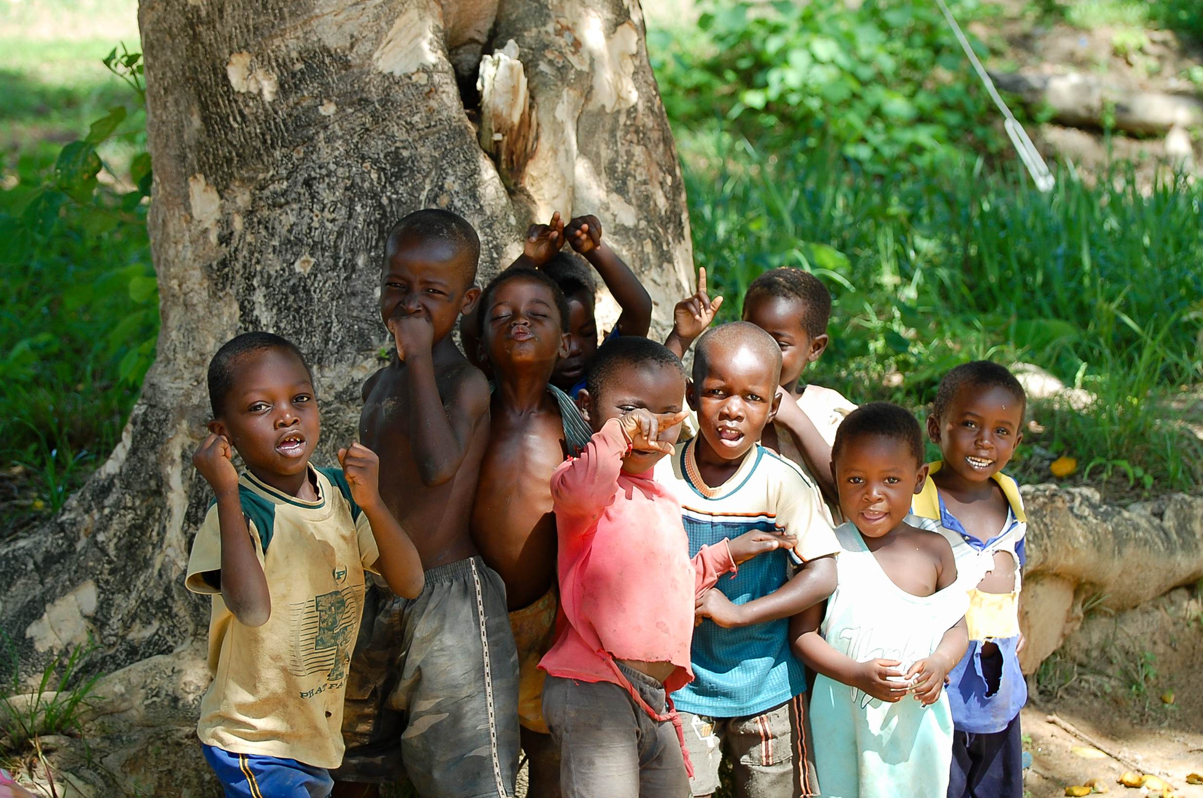 A group of children standing together under a tree, smiling and making playful gestures.