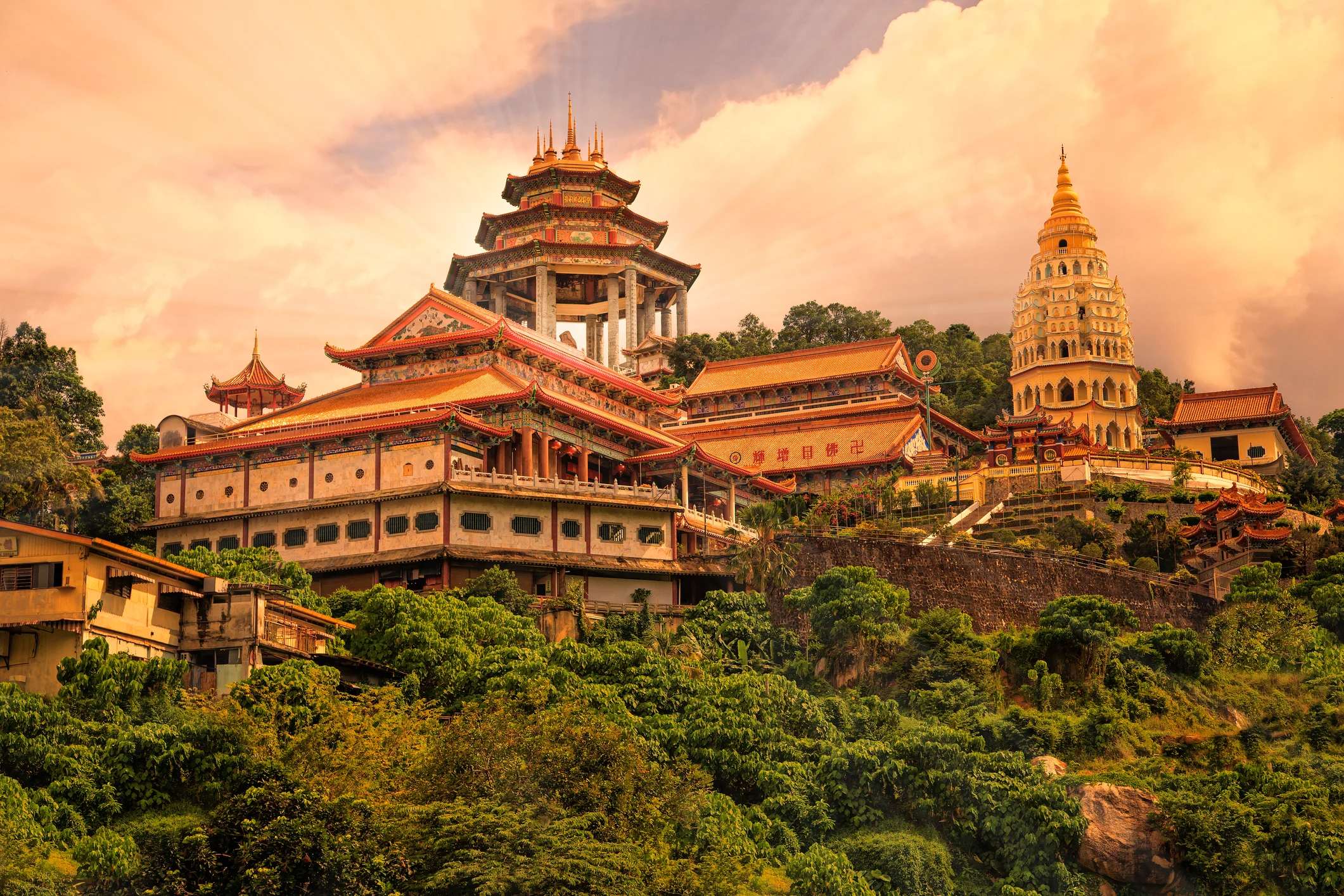 A scenic view of a majestic temple complex surrounded by lush greenery, with hills and a cloudy sky in the background.