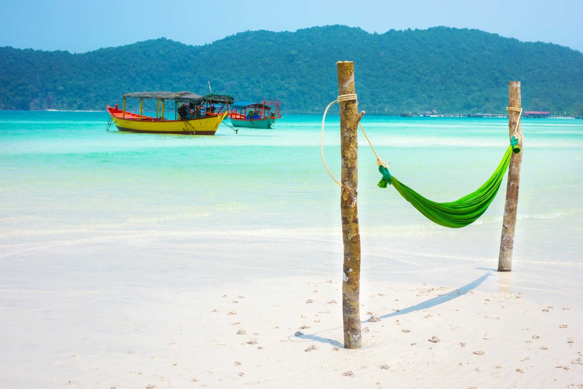 A green hammock between two wooden posts on a sandy beach, with boats and hills in the background.