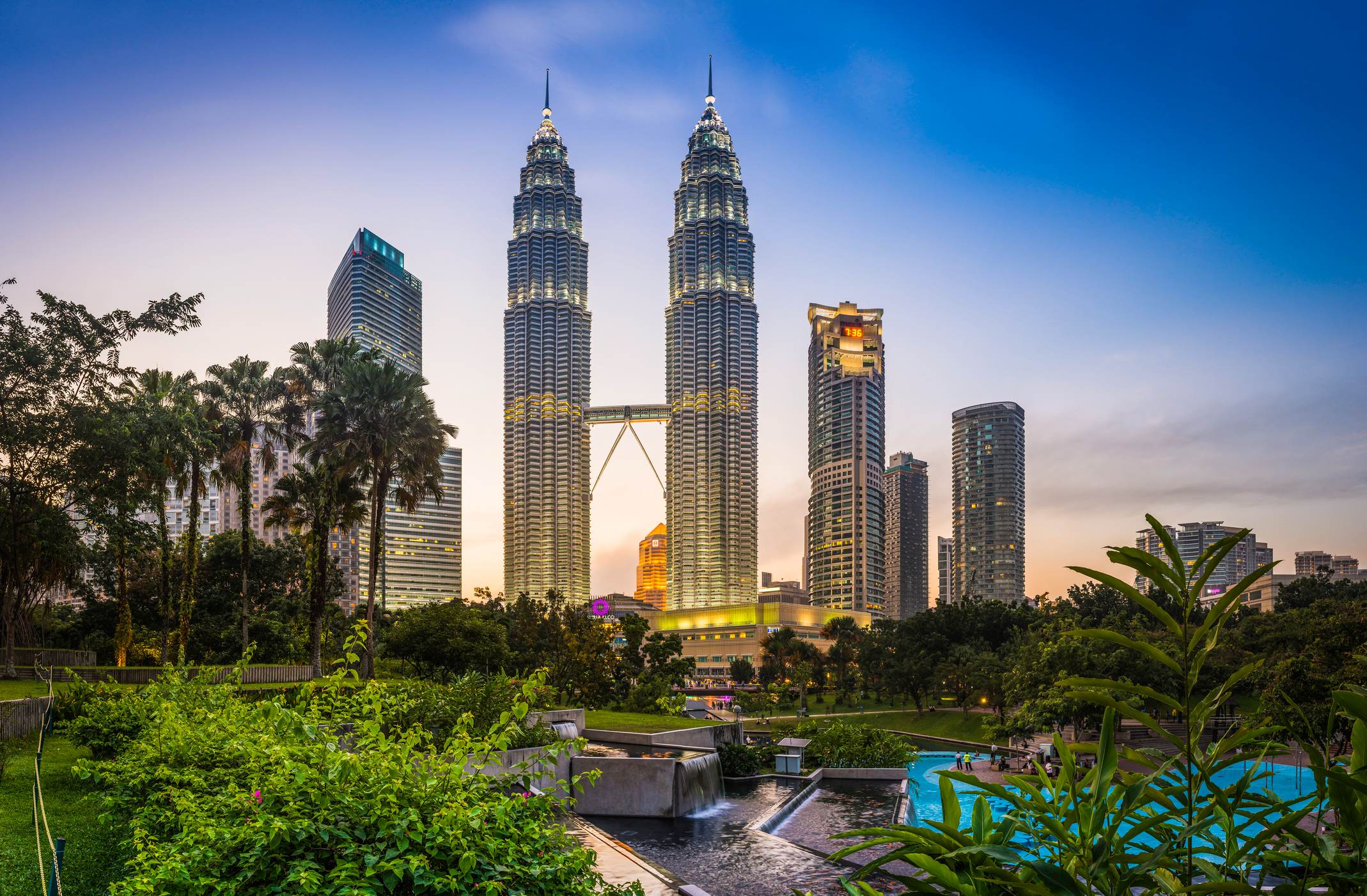 Petronas Towers rise above lush greenery at dusk, with city skyline in the background, showcasing modern architecture.