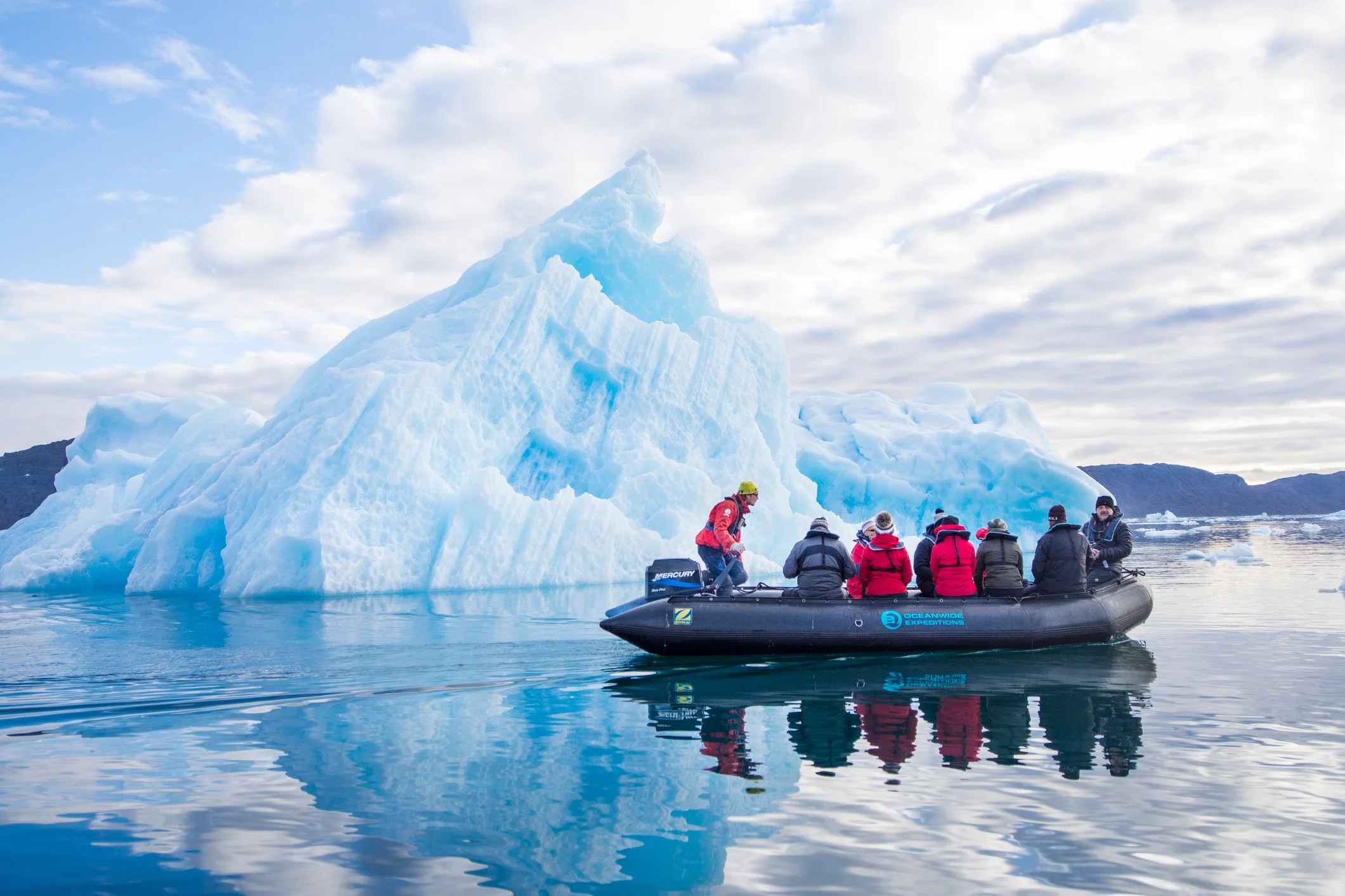A group of people in a boat near a large blue iceberg on calm water under a cloudy sky.