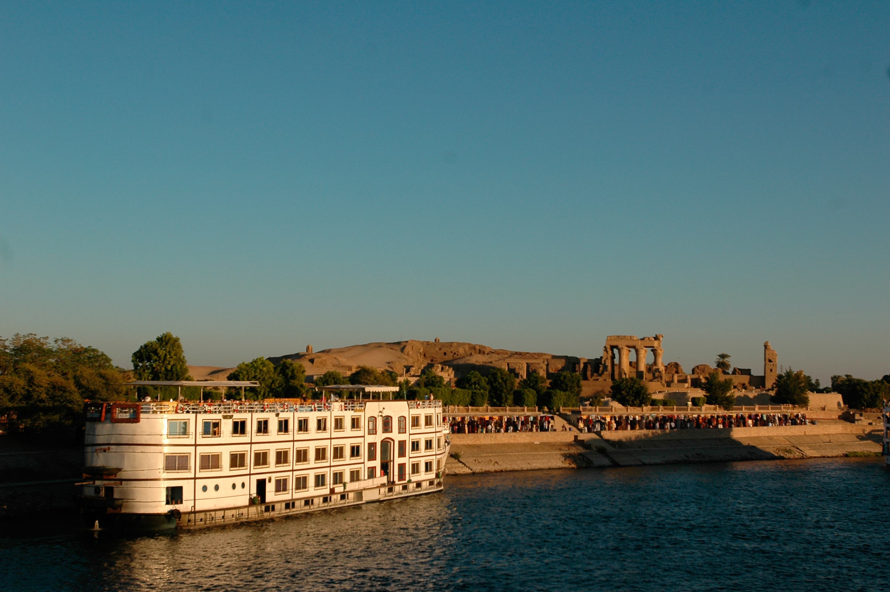 A riverboat anchored near ancient ruins, with clear blue skies and people gathered along the shoreline in the background.