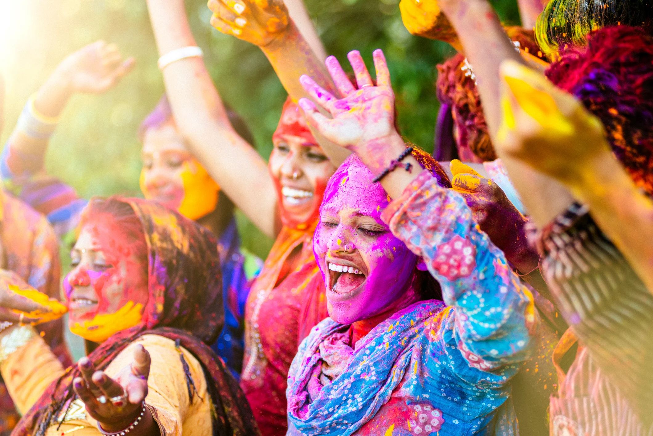 A group of joyful women celebrate with bright colors during a festival, smiling and raising their hands in excitement.