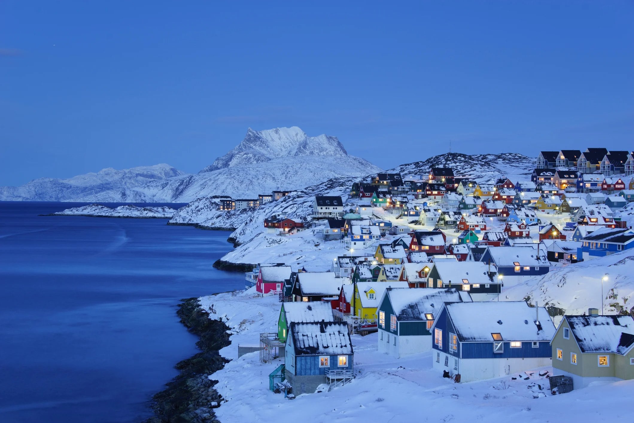 Colorful snowy houses line a coastal hill, with mountains in the background and a serene blue twilight sky.